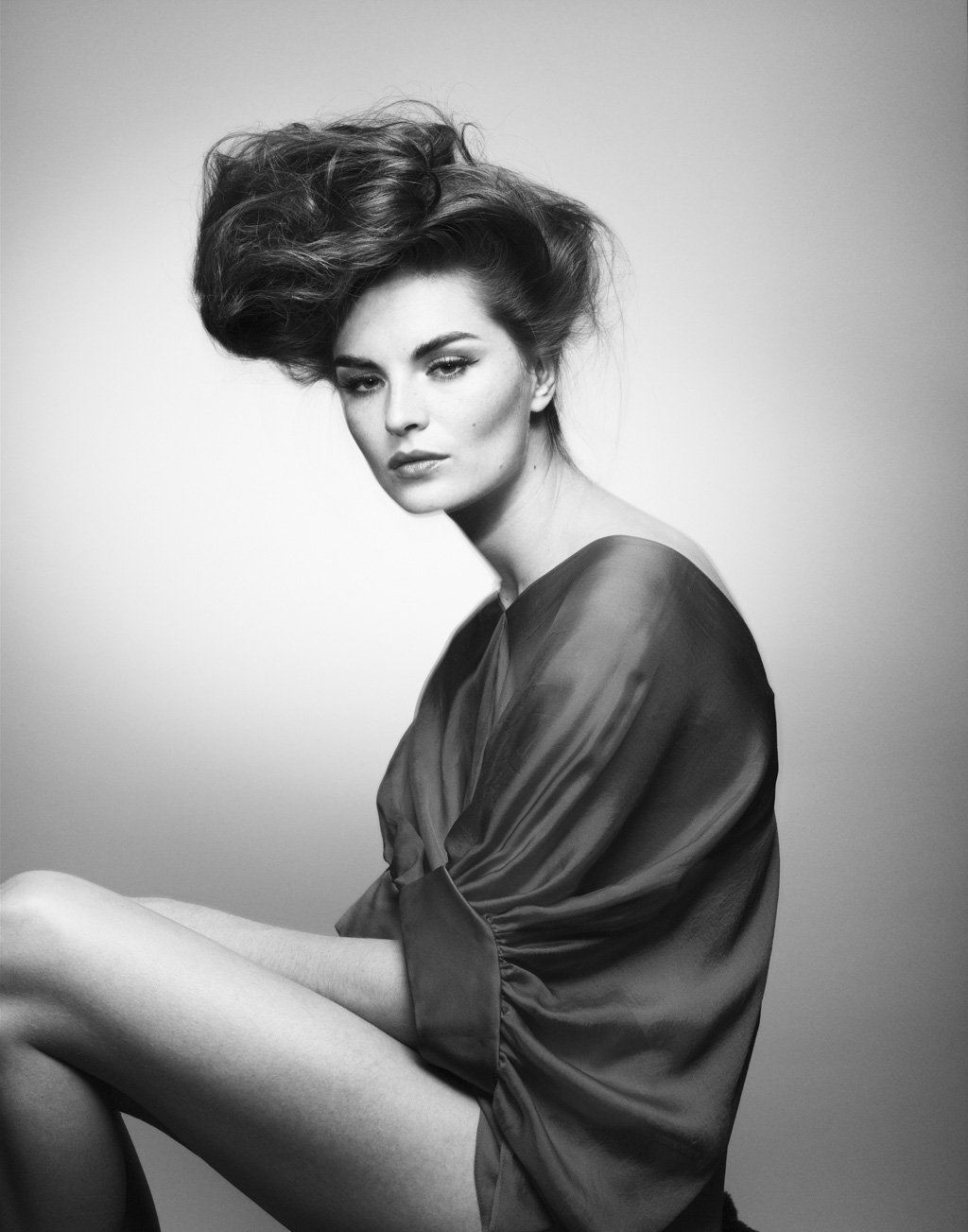 Black and white portrait of a woman with voluminous, styled hair, wearing a silky, loose garment, sitting with her knee slightly raised and looking directly at the camera.
