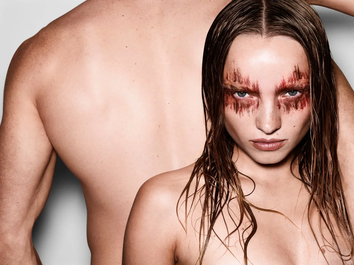 A woman with wet hair and intense makeup around her eyes, standing close to a shirtless man with light skin and freckles, in a close-up portrait.