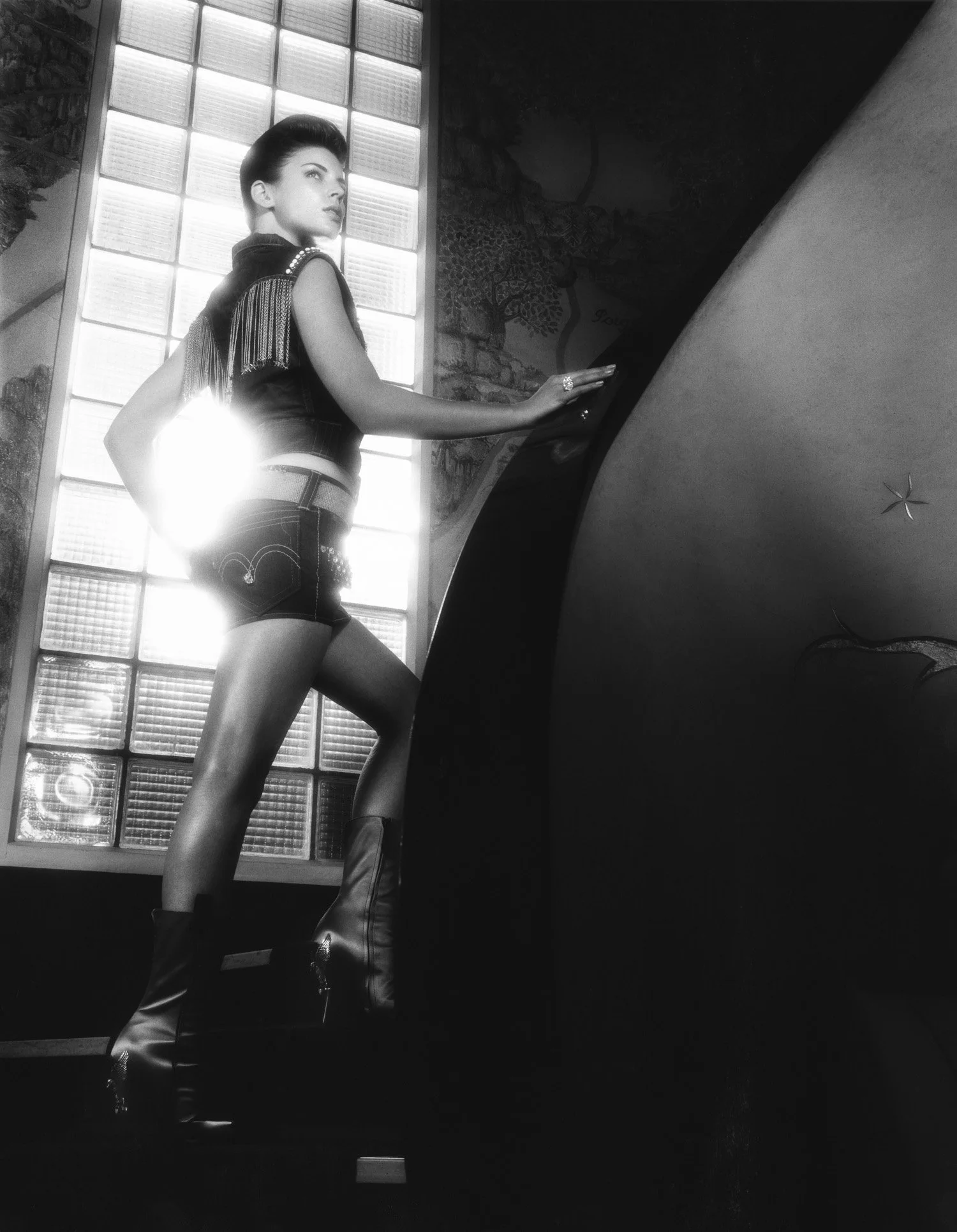 A woman with short dark hair standing at a piano, wearing a sleeveless top with fringe details, shorts, and high boots, in front of a large window with glass blocks.