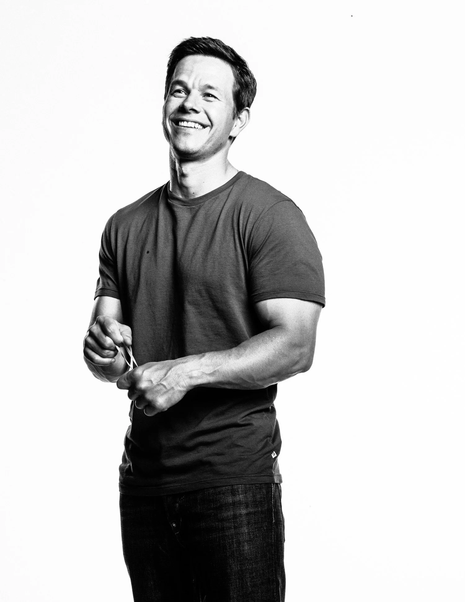 Black and white photo of MARK WAHLBERG with short dark hair, wearing a casual t-shirt and jeans, holding a pair of glasses in his hands.