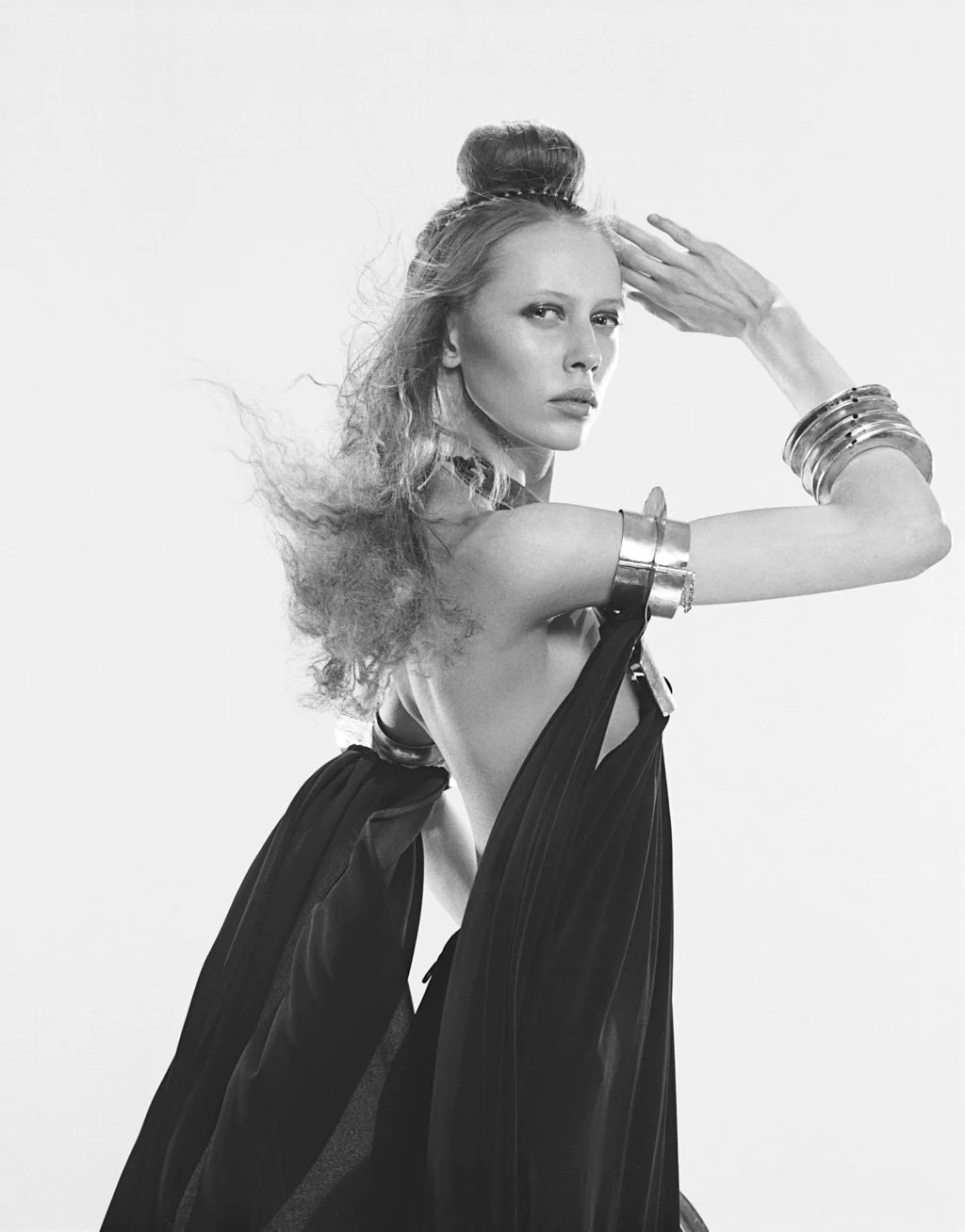 A black and white photo of a woman with wavy hair styled in an updo, wearing multiple metallic bangles on her arms, and a flowing, elegant black dress with a twisted design, posing with her hand on her forehead against a plain background.