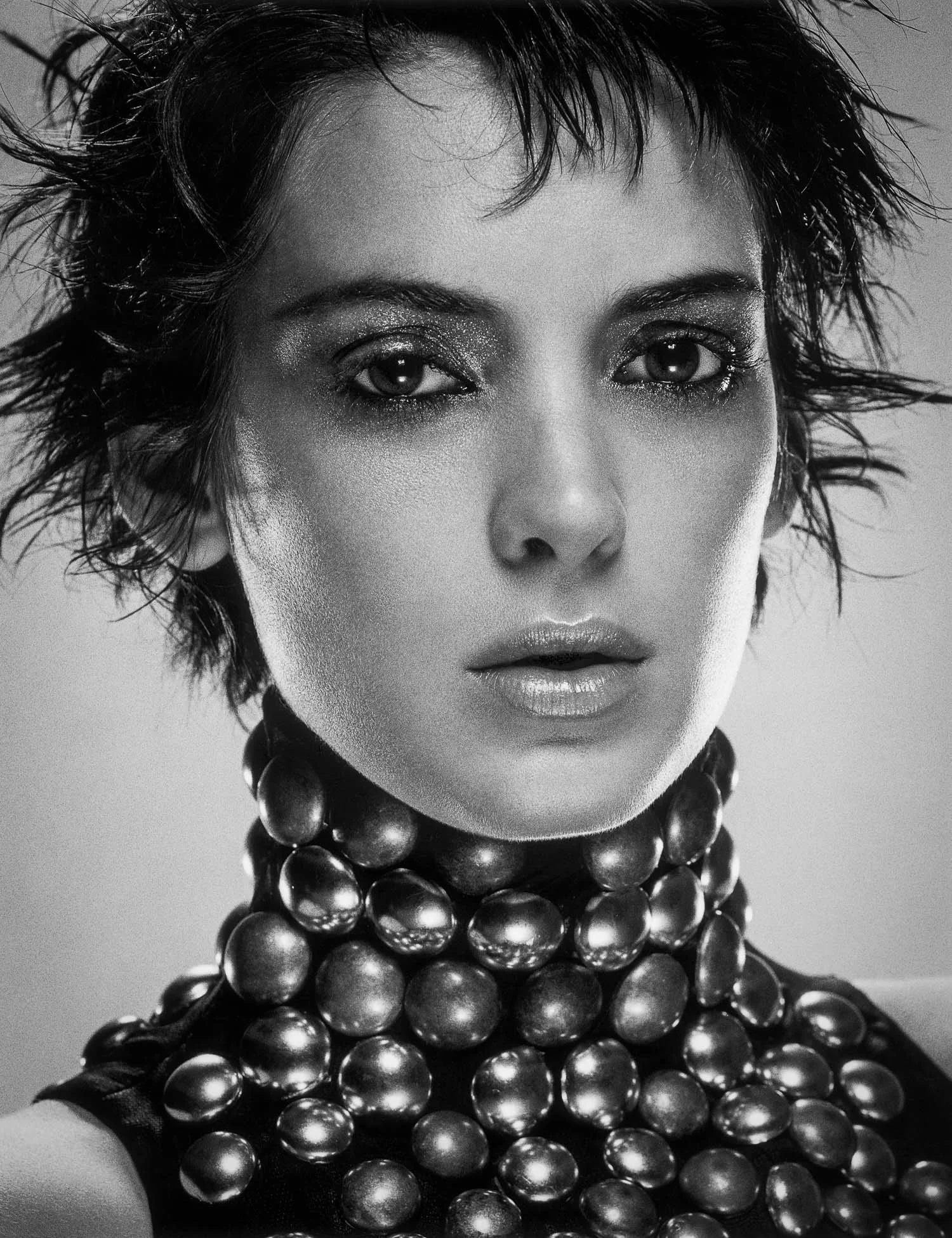 Black and white close-up portrait of WINONA RYDER with short, spiky hair wearing multiple strands of large, round beads around her neck.