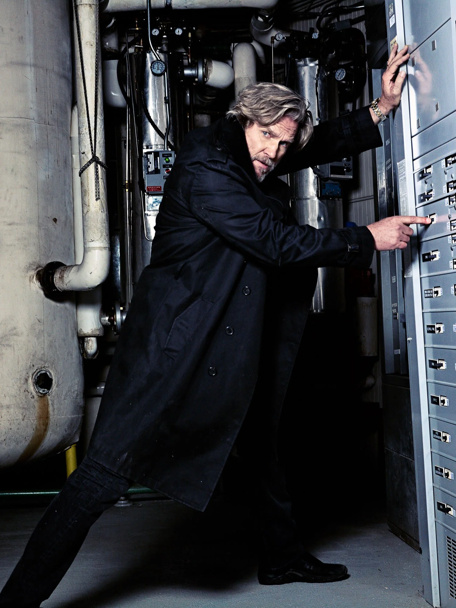 JEFF BRIDGES with gray hair and a beard is adjusting switches on an electrical panel in an industrial setting, wearing a black coat.