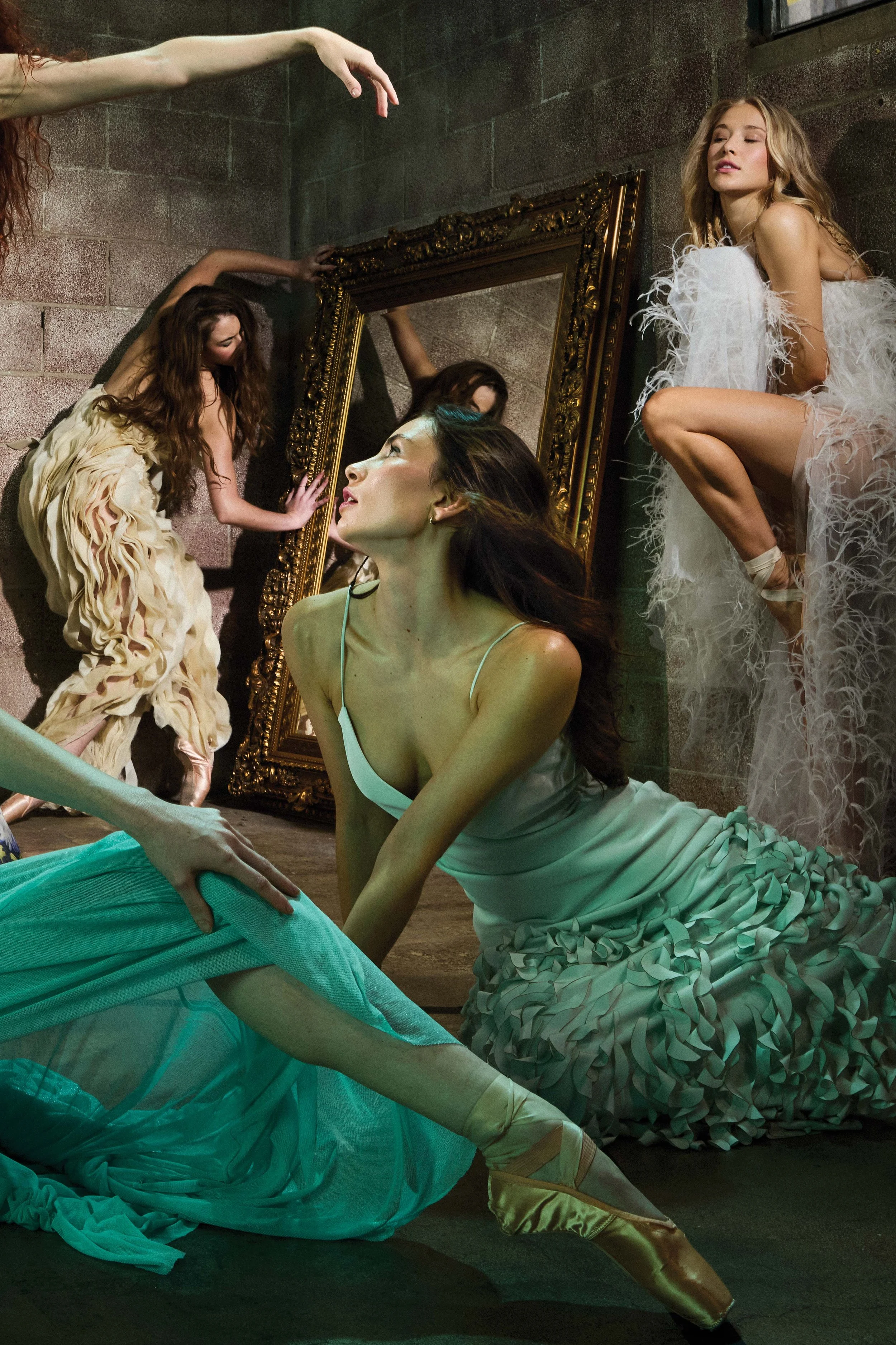Four women in ballet costumes are in a dance studio, with one woman sitting on the floor in the foreground, another quadruped with her back to the mirror, and two women in the background, one reaching up and the other squatting, all surrounded by a l