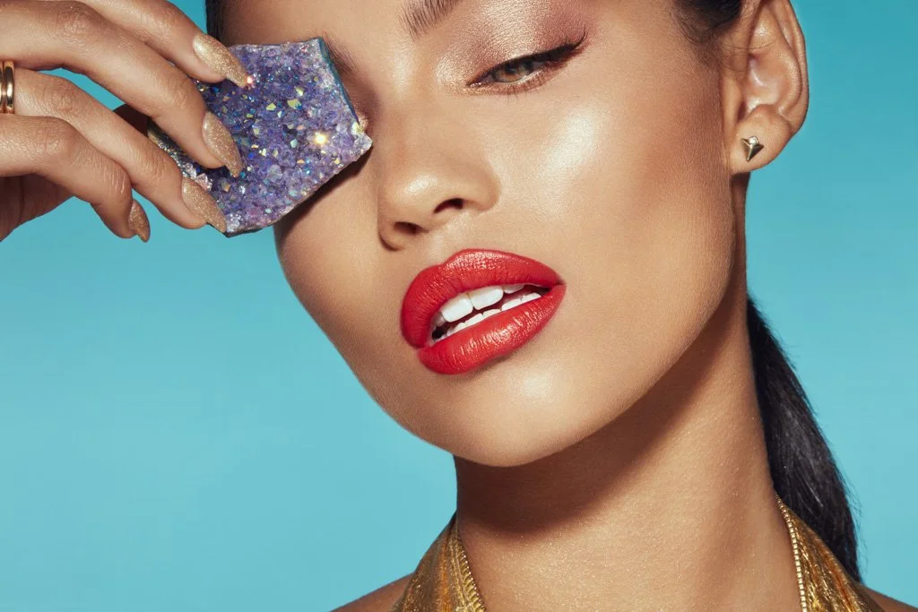 A woman with red lipstick is holding a sparkly purple cloth near her eye against a bright blue background.