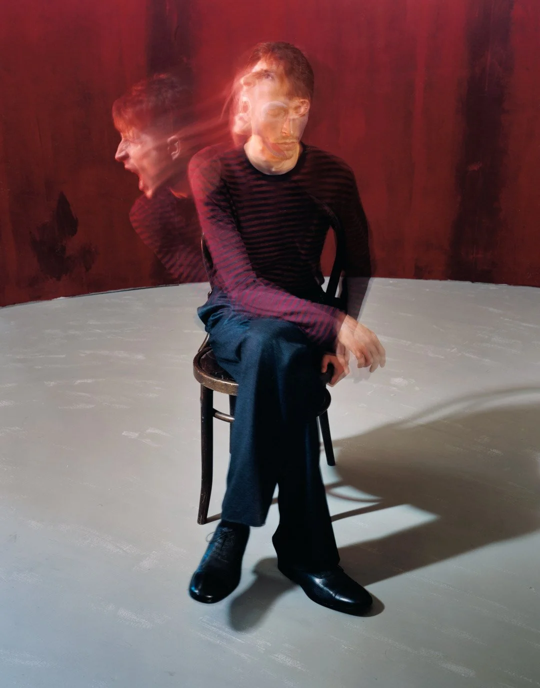 A man sitting on a chair, with a motion blur effect creating two overlapping images of him, set against a red wall and gray floor.