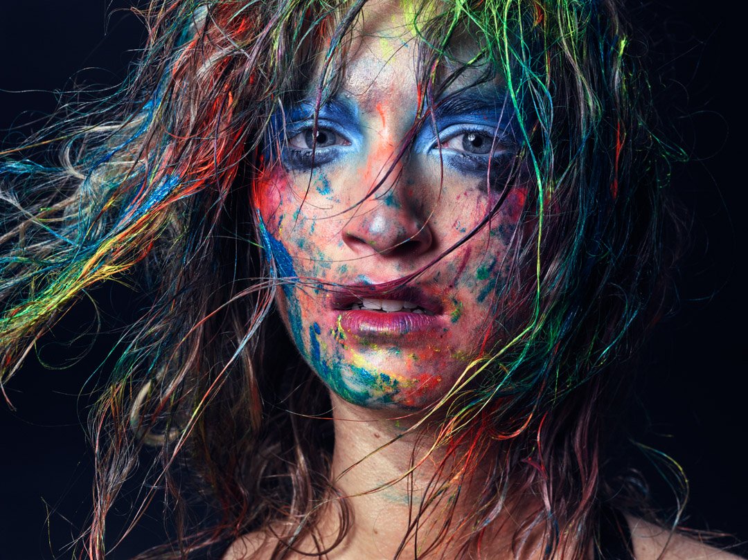 Close-up of a woman with messy, wet hair covered in colorful paint splatters on her face and neck, against a black background.