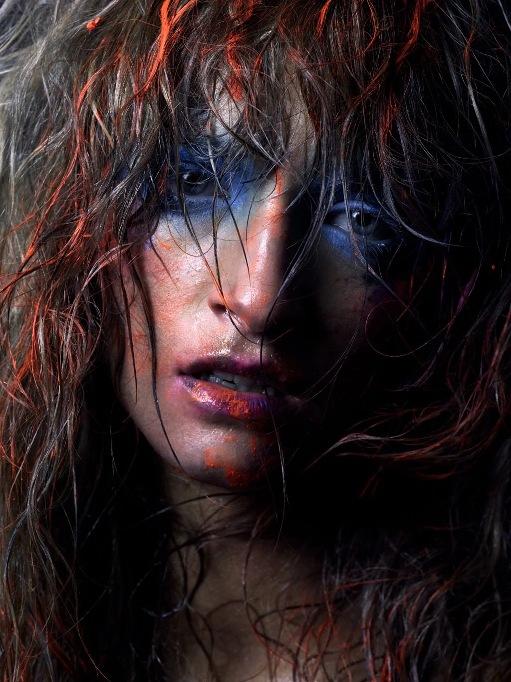 Close-up of a woman with wet, reddish-brown hair partially covering her face, with intense makeup including blue and red accents around her eyes and lips.