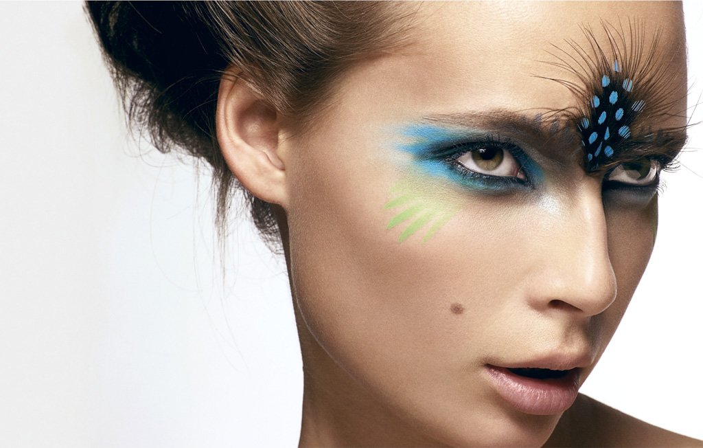 Close-up of a woman with creative makeup, featuring blue and green accents around her eyes and a decorative black and blue feather accessory on her eyebrow.