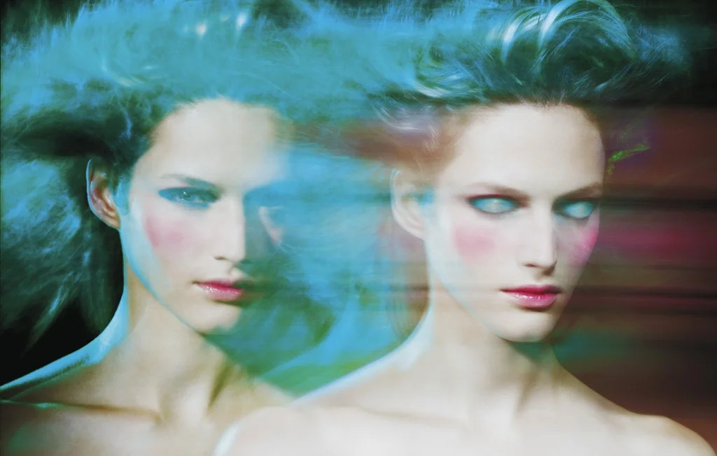 Two women with makeup and styled hair, with a colorful, blurred background, one looking directly at the camera and the other with eyes slightly closed.