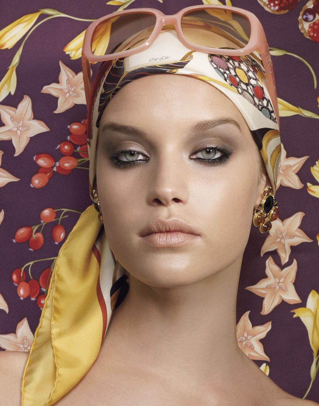 Portrait of a woman with blonde hair, wearing a patterned headscarf and pink sunglasses on her forehead, lying on a floral and berry-patterned background, with gold earrings and makeup.