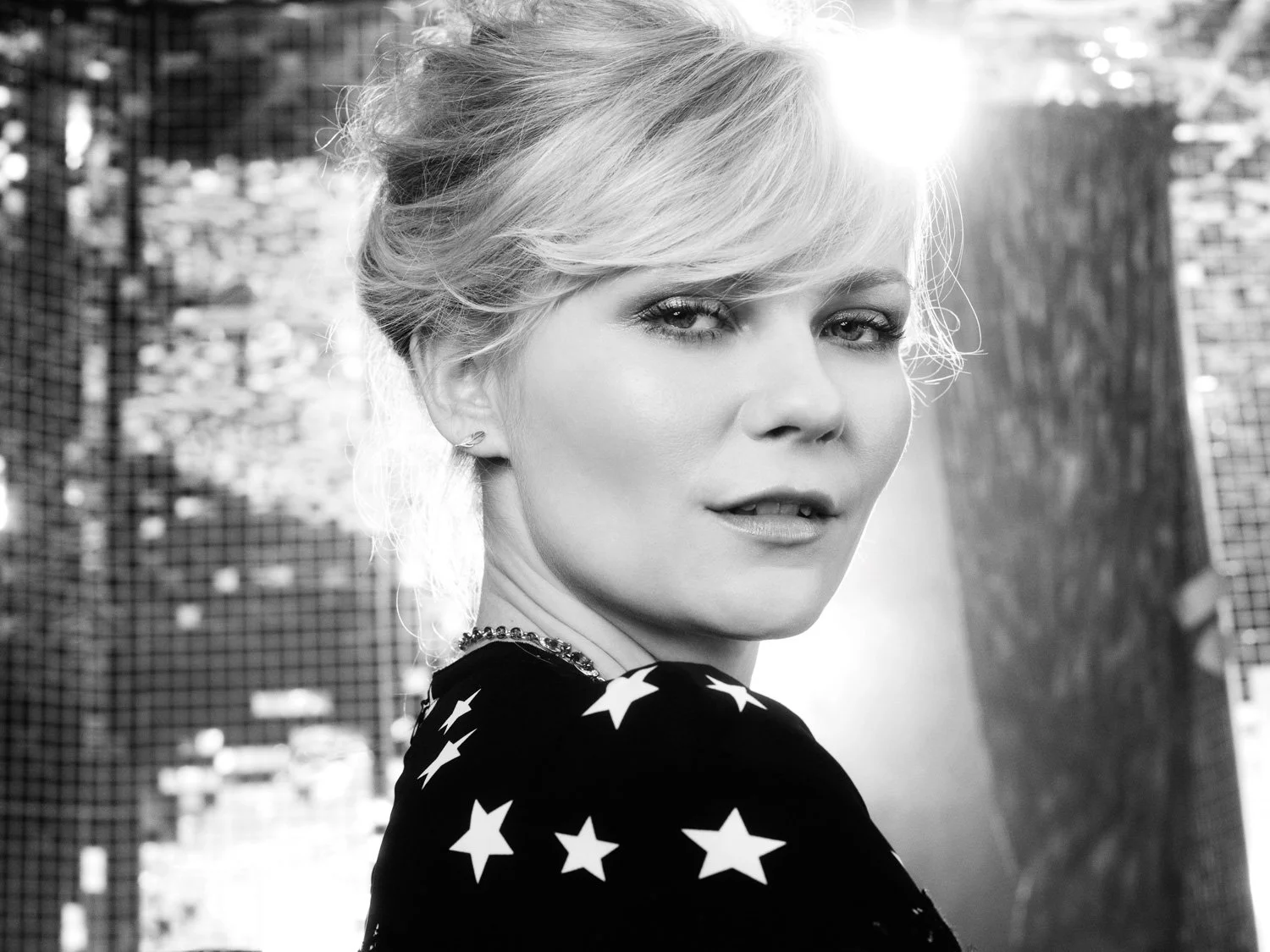 Black and white portrait of KIRSTEN DUNST with light-colored hair styled in an updo, wearing a star-patterned garment, with sunlight behind her.