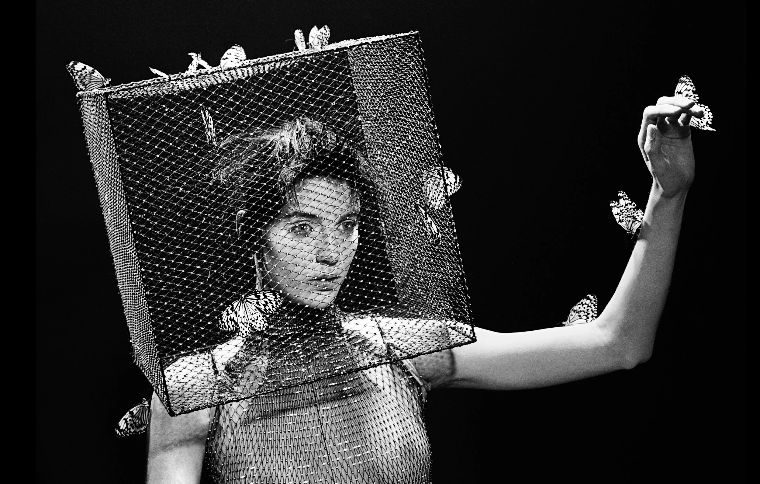 A woman with dark hair and a patterned top, wearing a mesh cube hat adorned with butterflies, with matching butterflies flying around her in a black and white photograph.
