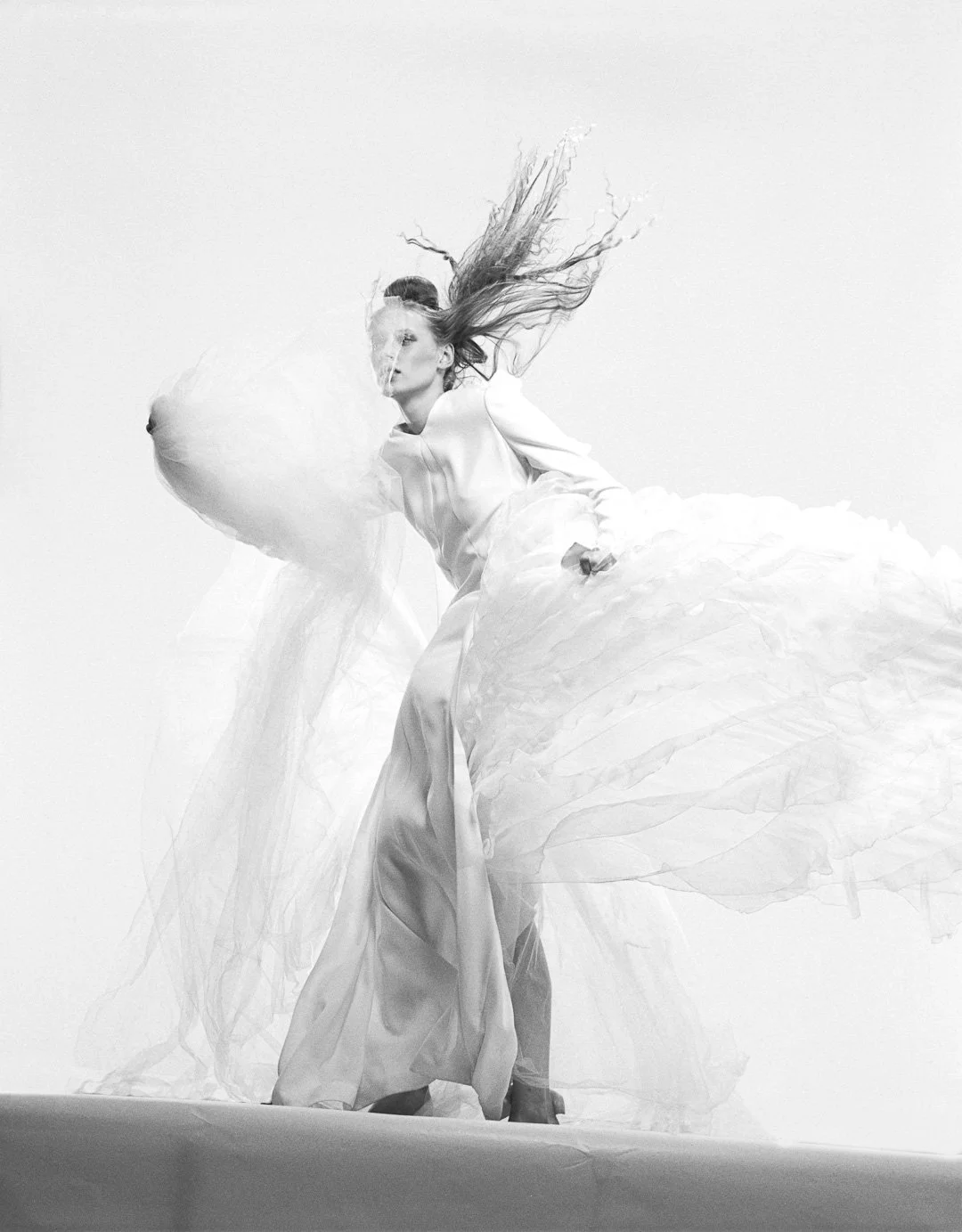 Black and white photo of a woman in flowing dress with long hair, posing dramatically against a plain background.