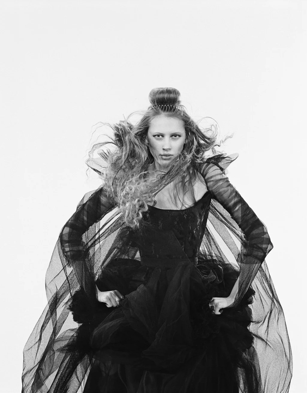 A woman with long, curly hair in a high bun, wearing a black lace dress with puffed, sheer sleeves, sitting with hands on her hips against a plain white background.