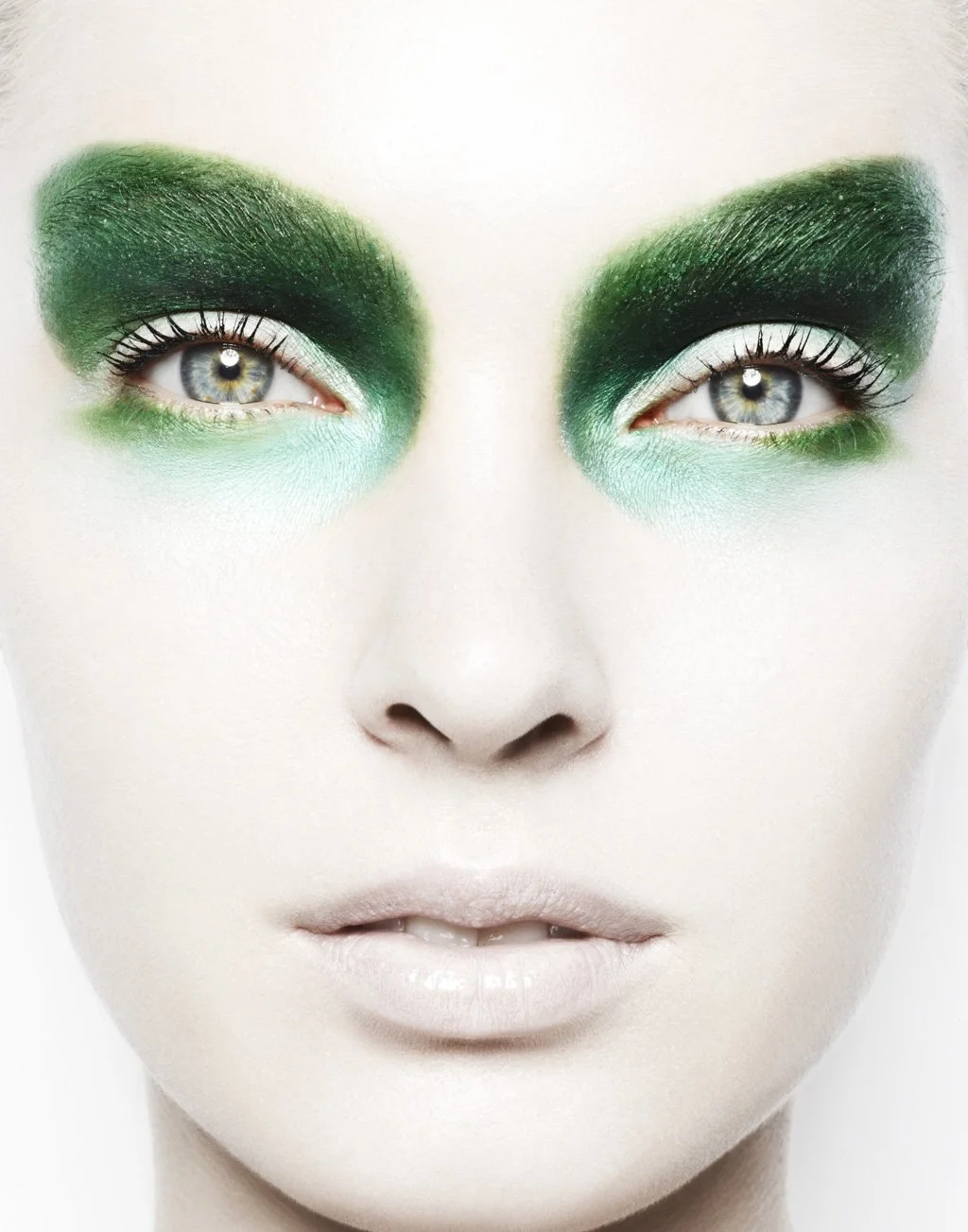 Close-up of a person's face with striking makeup featuring vibrant green eyeshadow and light skin, with the person's eyes wide open and lips slightly parted.