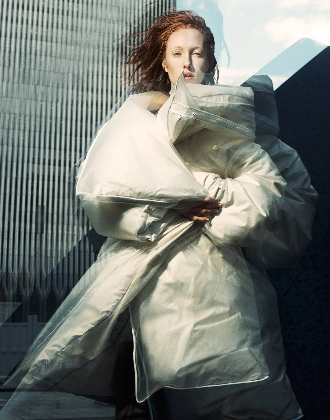 A woman with red hair standing behind a glass window, holding a large, light-colored quilted coat or garment, with modern buildings and a cloudy sky in the background.