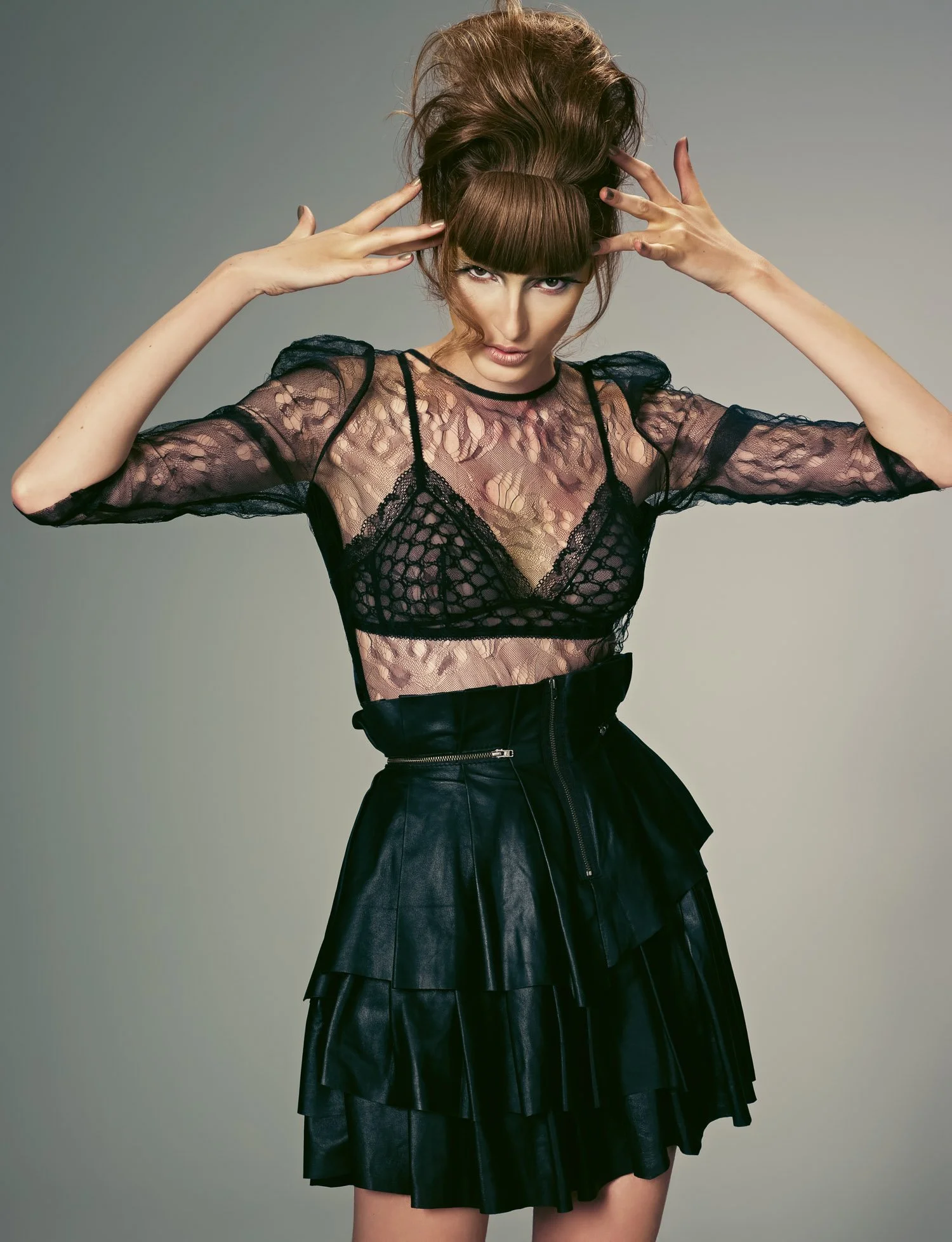 Fashion model with styled hair and bangs wearing a black lace top and black leather skirt posing with hands near her head.
