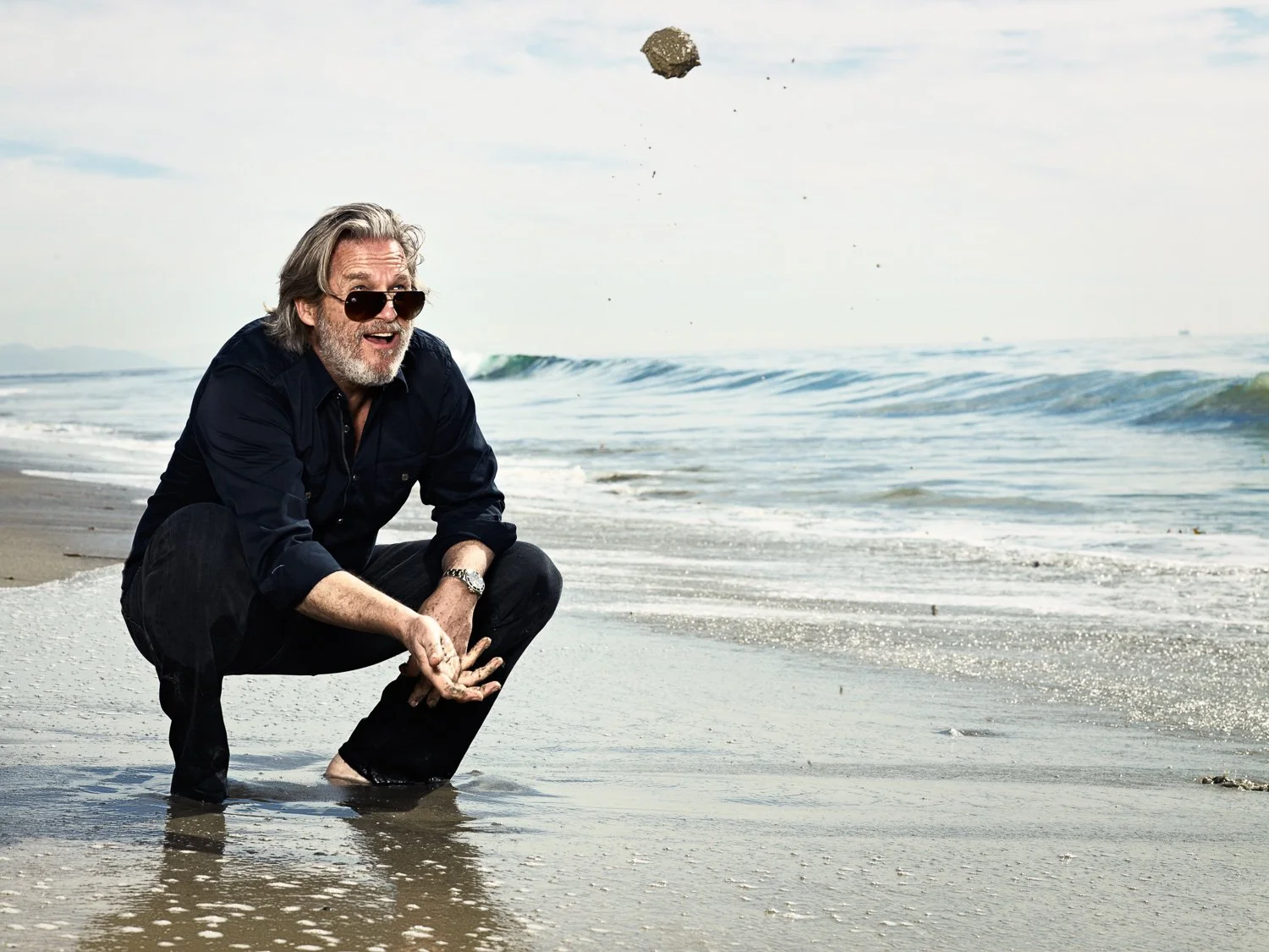 JEFF BRIDGES with gray hair and beard, wearing sunglasses, a dark shirt, and dark pants, squatting on a beach with his feet in shallow water, as a large rock falls from the sky above him.