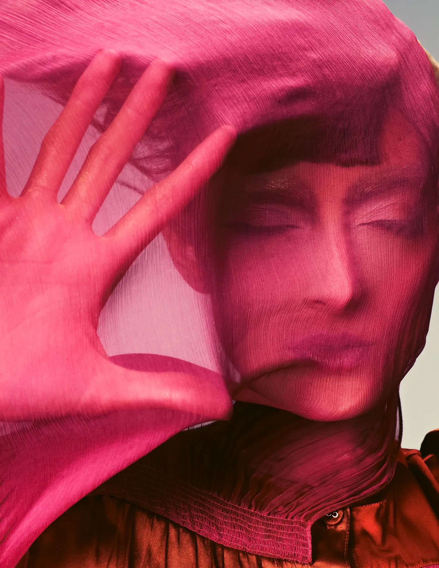 A person wearing a pink sheer fabric over their face and head, with their eyes closed, holding their hand up near their face, situated behind the fabric.