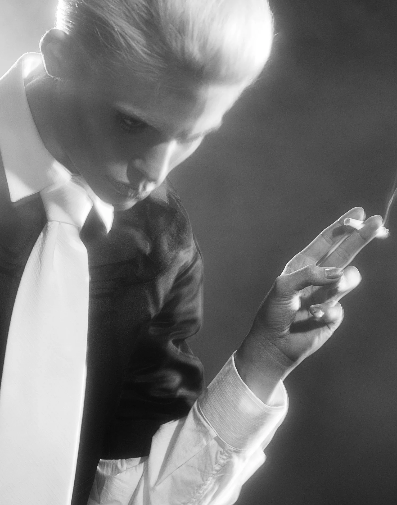 A black and white photo of a woman with platinum blonde hair tied back, wearing a white shirt, black leather jacket, and white tie. She is looking down, holding a lit cigarette in her hand.