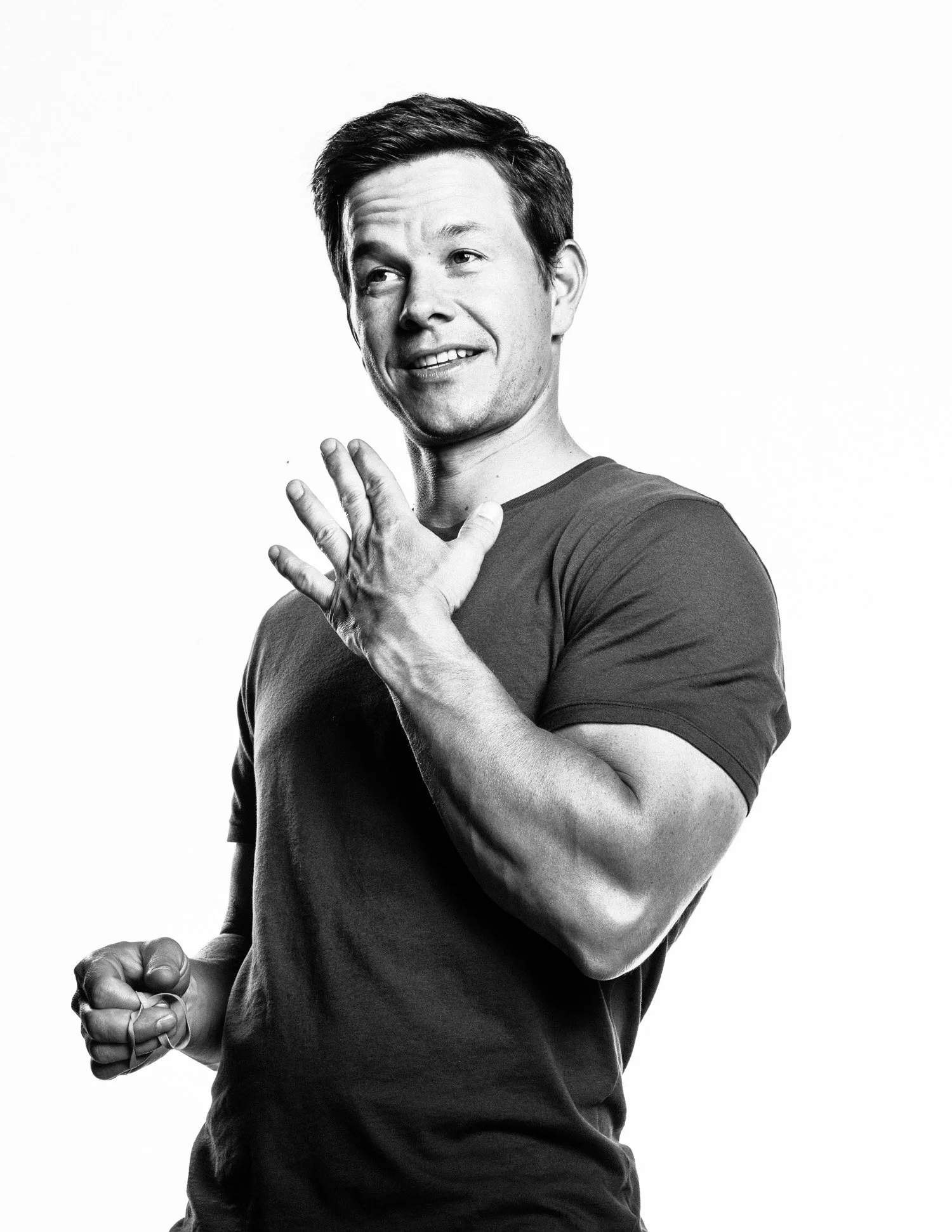 A black and white photo of MARK WAHLBERG with short hair, smiling, wearing a dark t-shirt, and making a gesture with his hand on his chest.
