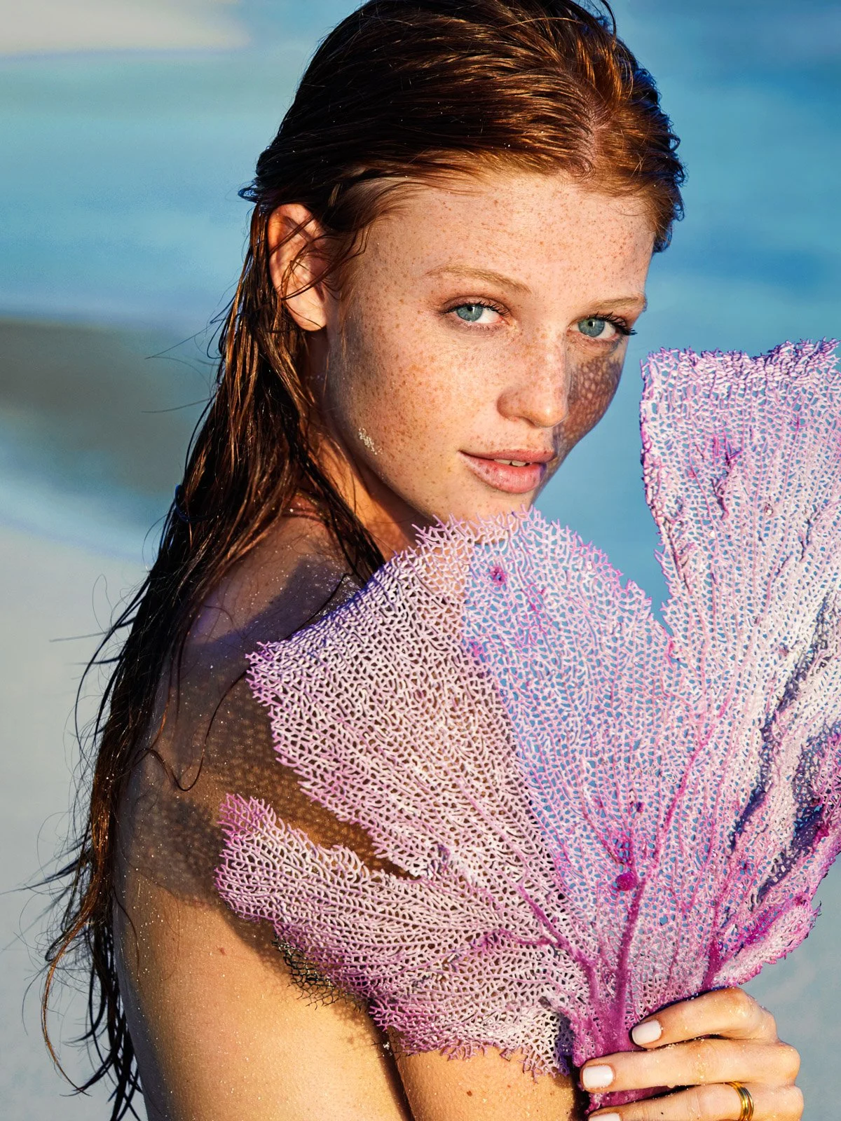 A woman with wet reddish-brown hair and freckles on her face holding a large pink and purple sea fan coral at the beach.
