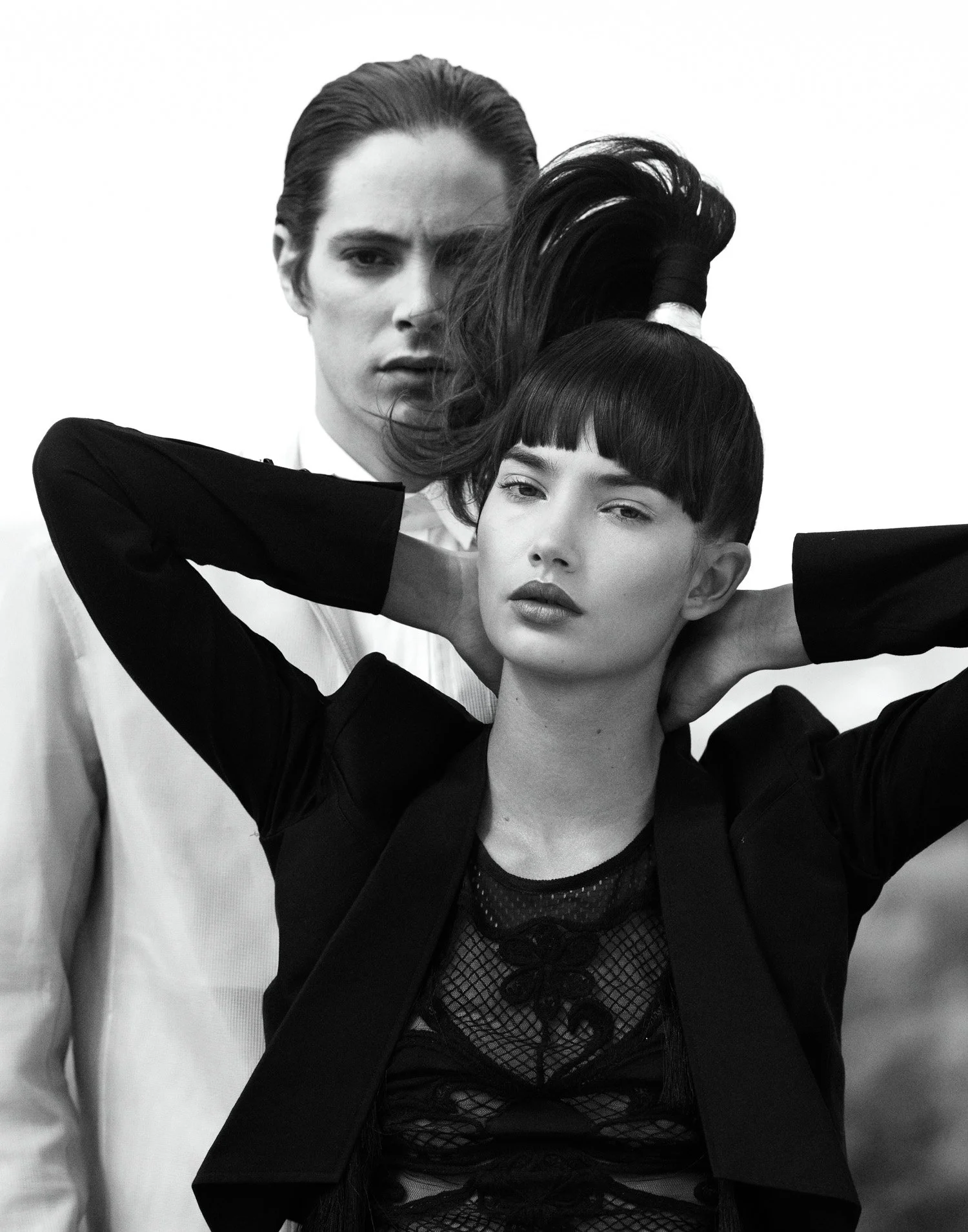 Black and white photo of two women, one with short black hair and the other with slicked back hair, posing with serious expressions, arms behind their heads, against a plain background.