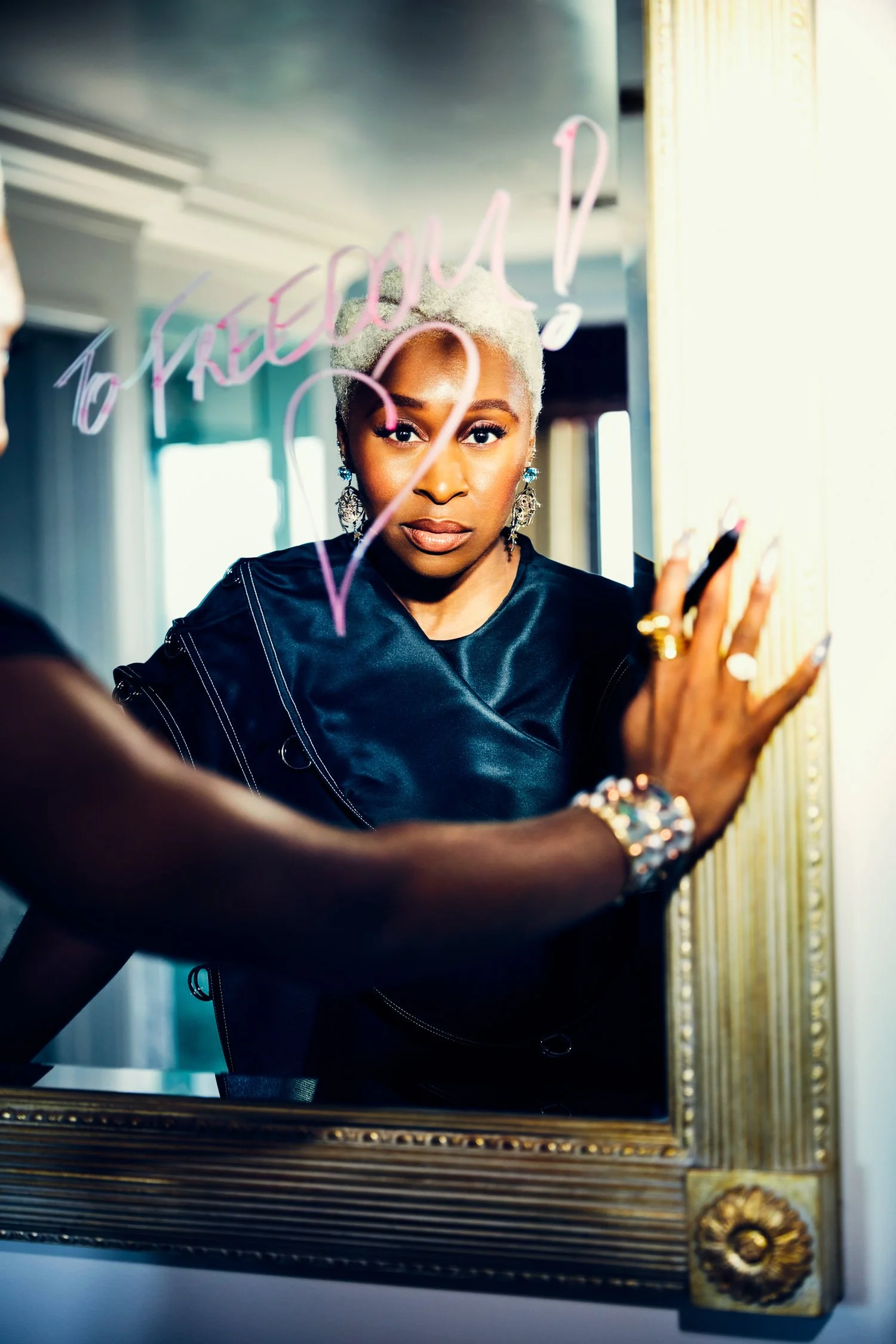 A woman with platinum blonde hair and earrings looks into a mirror, writing a message on it with a pink marker.