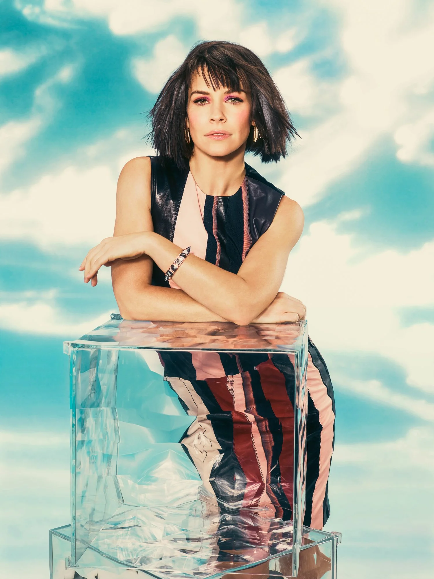 EVANGELINE LILLY with dark, shoulder-length hair and pink makeup, wearing a sleeveless black leather top and patterned pants, leaning on a transparent acrylic table with sky and clouds in the background.