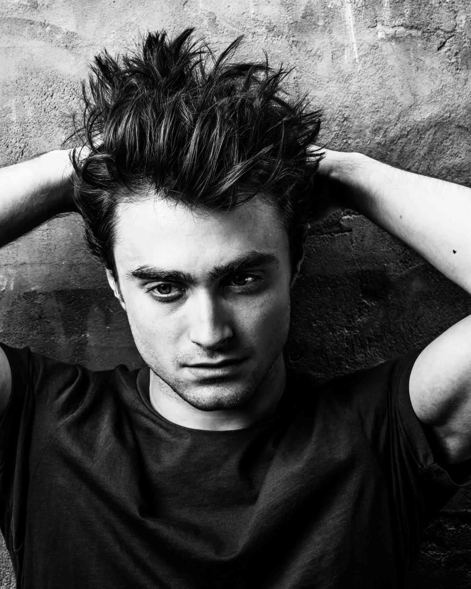 Black and white photo of DANIEL RADCLIFFE with tousled hair, wearing a dark T-shirt, lying against a textured wall with arms behind his head.
