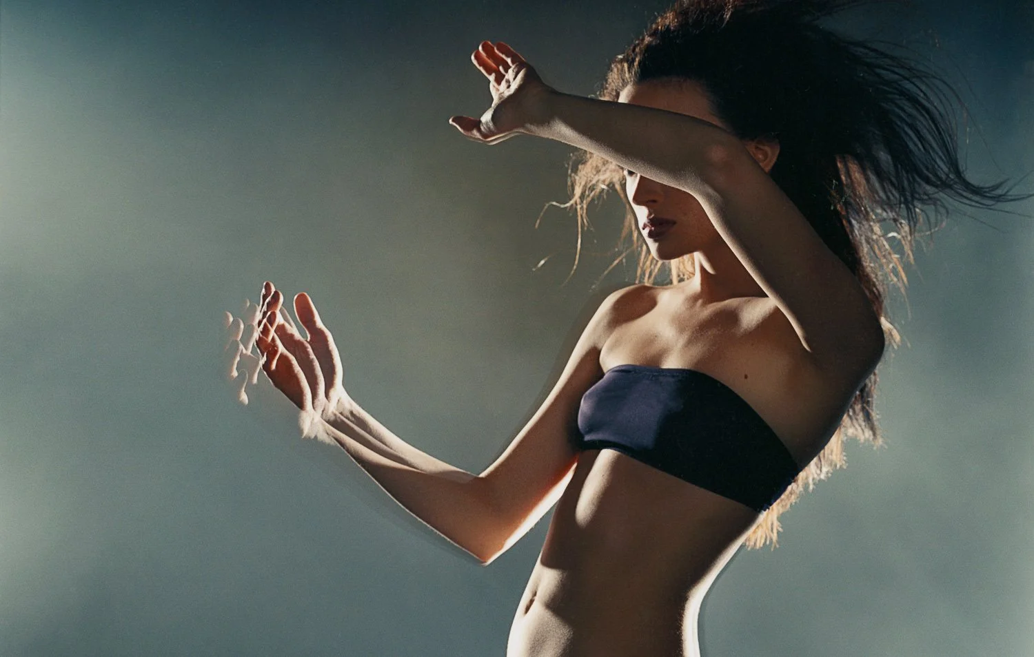 A woman with long hair and light skin wearing a black strapless top, posing with her arms raised, against a subdued background with soft lighting.