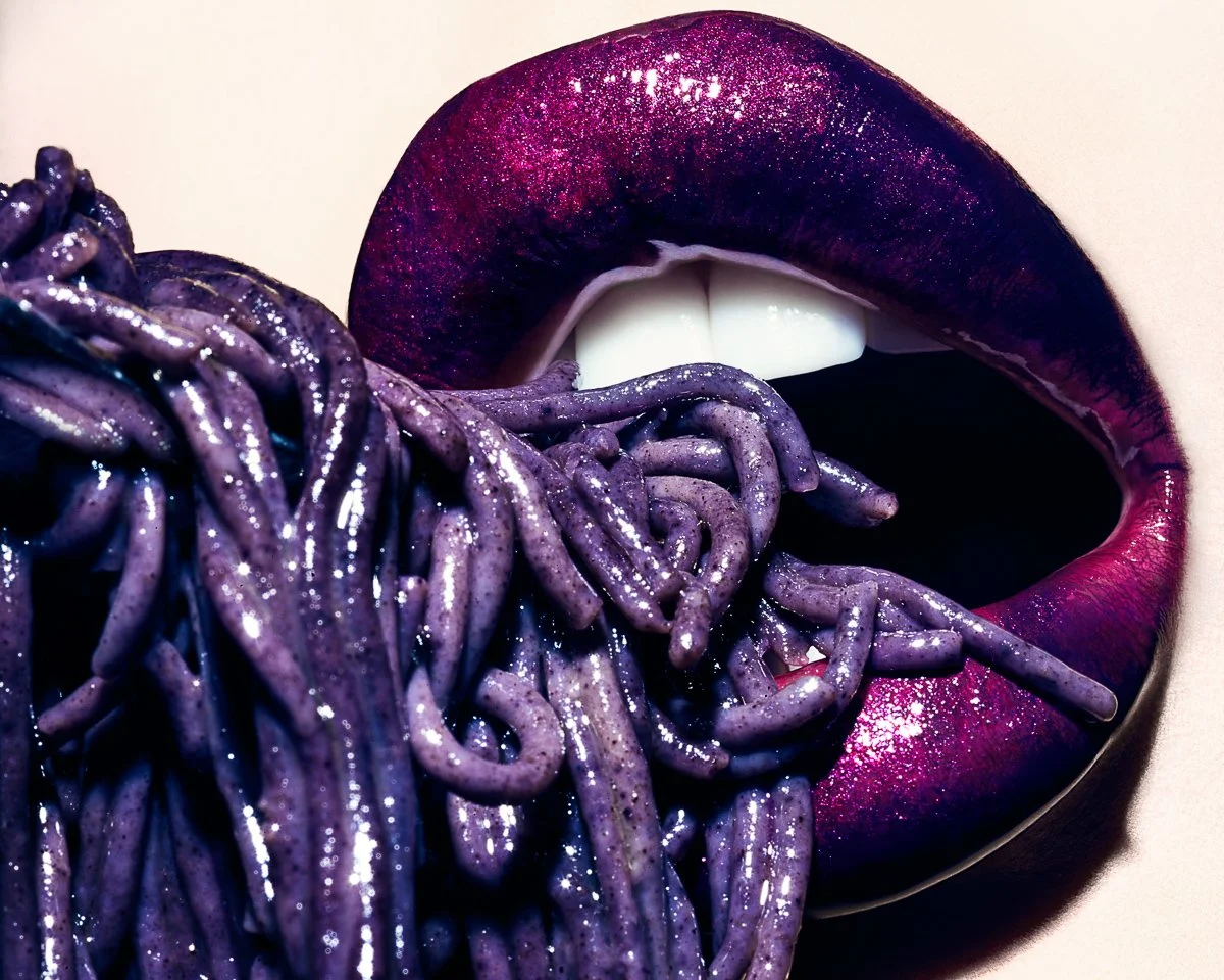 Close-up of a person with metallic purple lipstick and purple lipstick, with purple spaghetti noodles in their mouth.