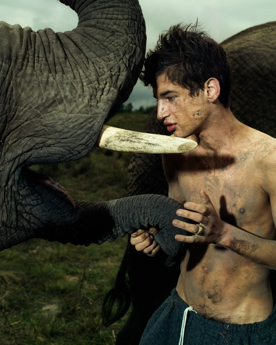 A shirtless man with dirt on his face and body holds an elephant's trunk while an elephant touches him with its trunk, in an outdoor grassy area.