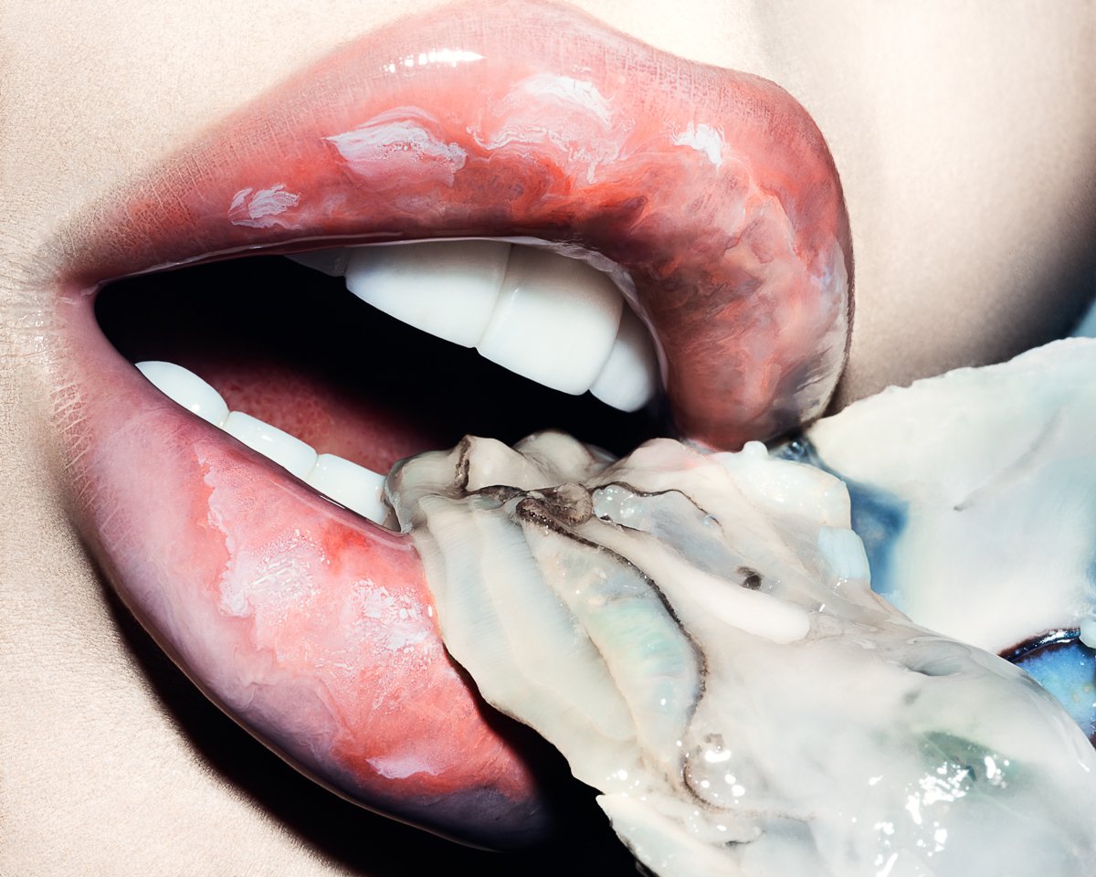 A close-up of a person's mouth with red lipstick, slightly open, with a piece of raw oyster being held by a hand near the lips.