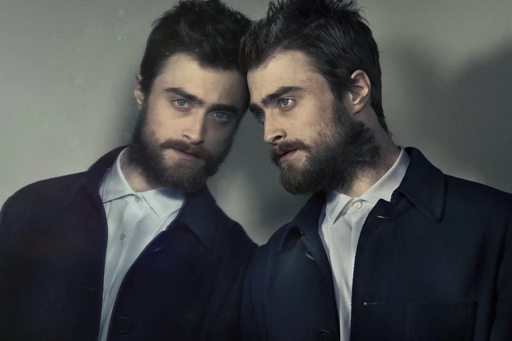 DANIEL RADCLIFFE with beards and dark hair leaning closely against each other, looking intently at the camera, wearing dark jackets and white shirts.