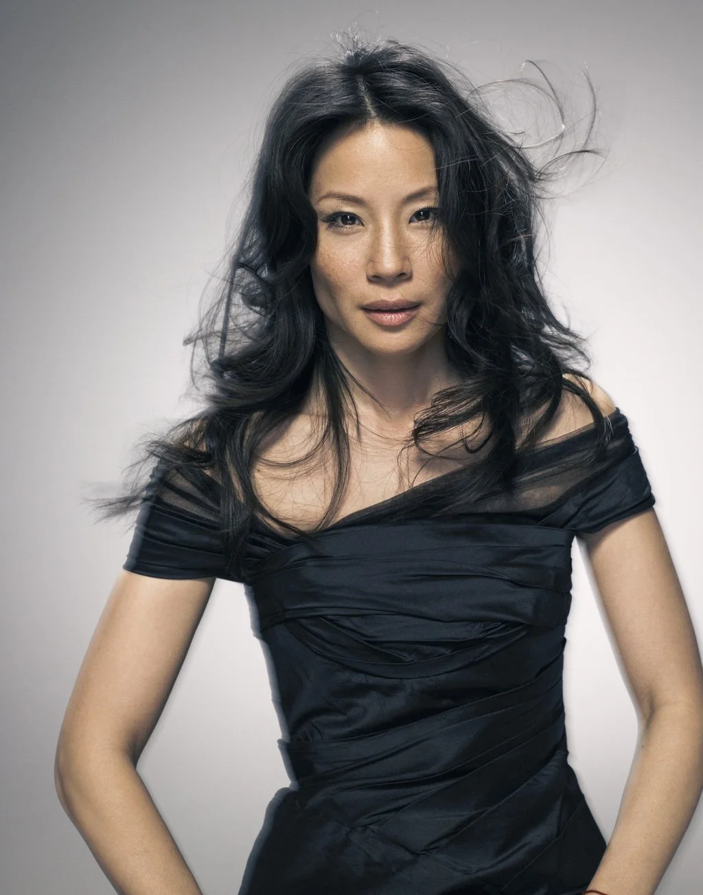 LUCY LIU with long, wavy black hair and light skin poses against a plain background, wearing an off-shoulder black dress.