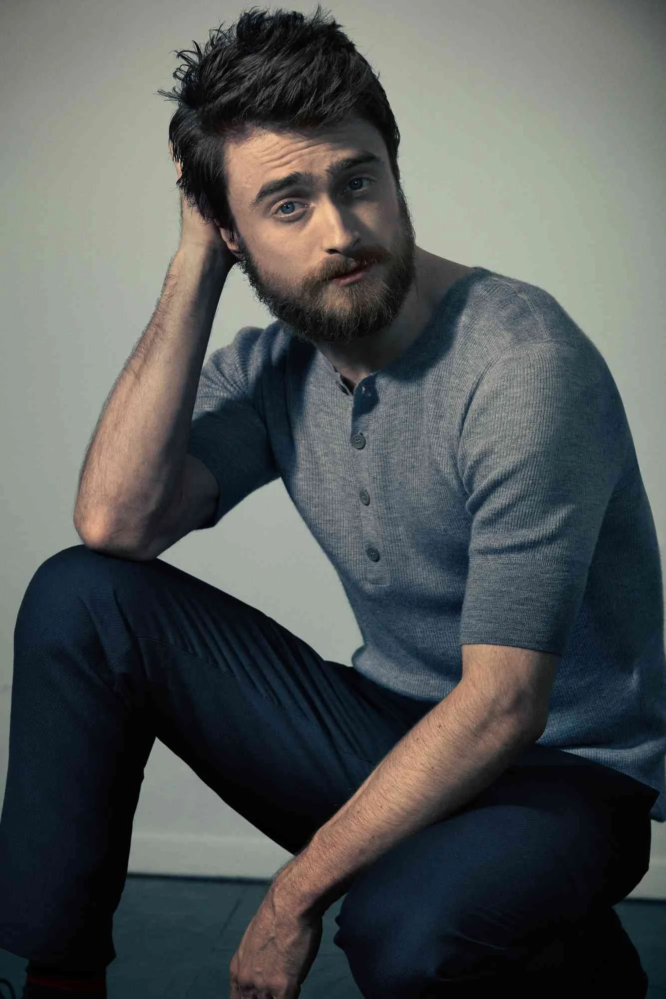 DANIEL RADCLIFFE with dark hair and a beard, wearing a gray, button-up shirt, seated against a plain background. He rests one arm on his knee and touches the back of his head with his other hand, looking directly at the camera.