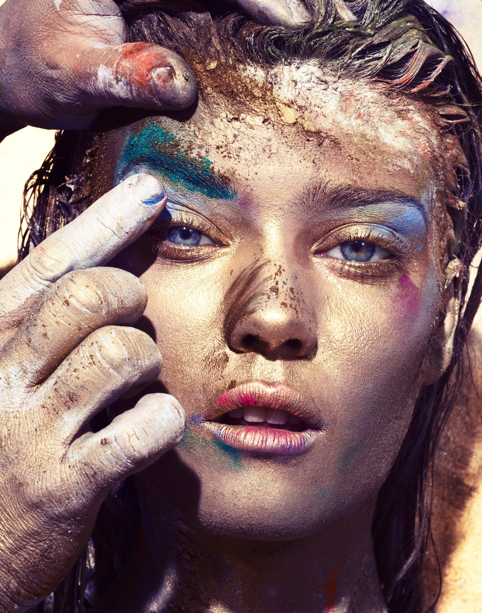 Close-up of a woman with colorful paint on her face and hands, with wet hair and bright blue eyes.