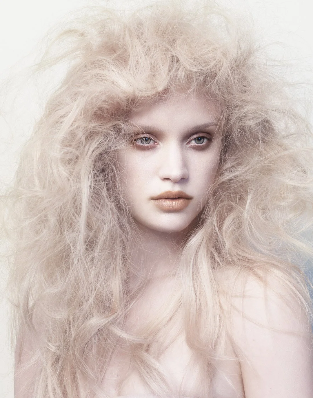 Close-up portrait of a woman with very light blonde, voluminous, curly hair and pale skin, looking directly at the camera.