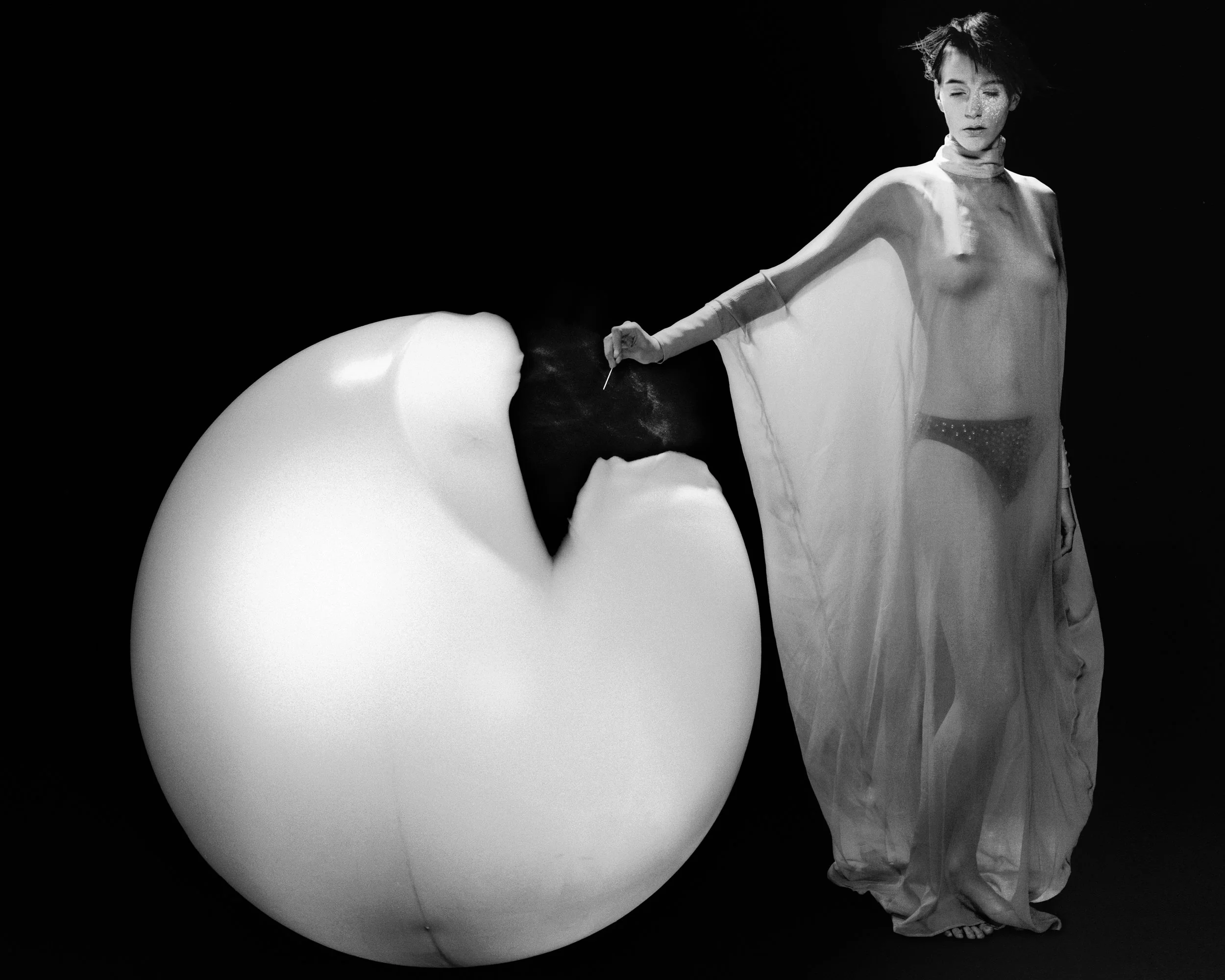 A woman in sheer, flowing clothing with short, tousled hair stands next to a large, cracked eggshell. The scene is in black and white with a dark background.