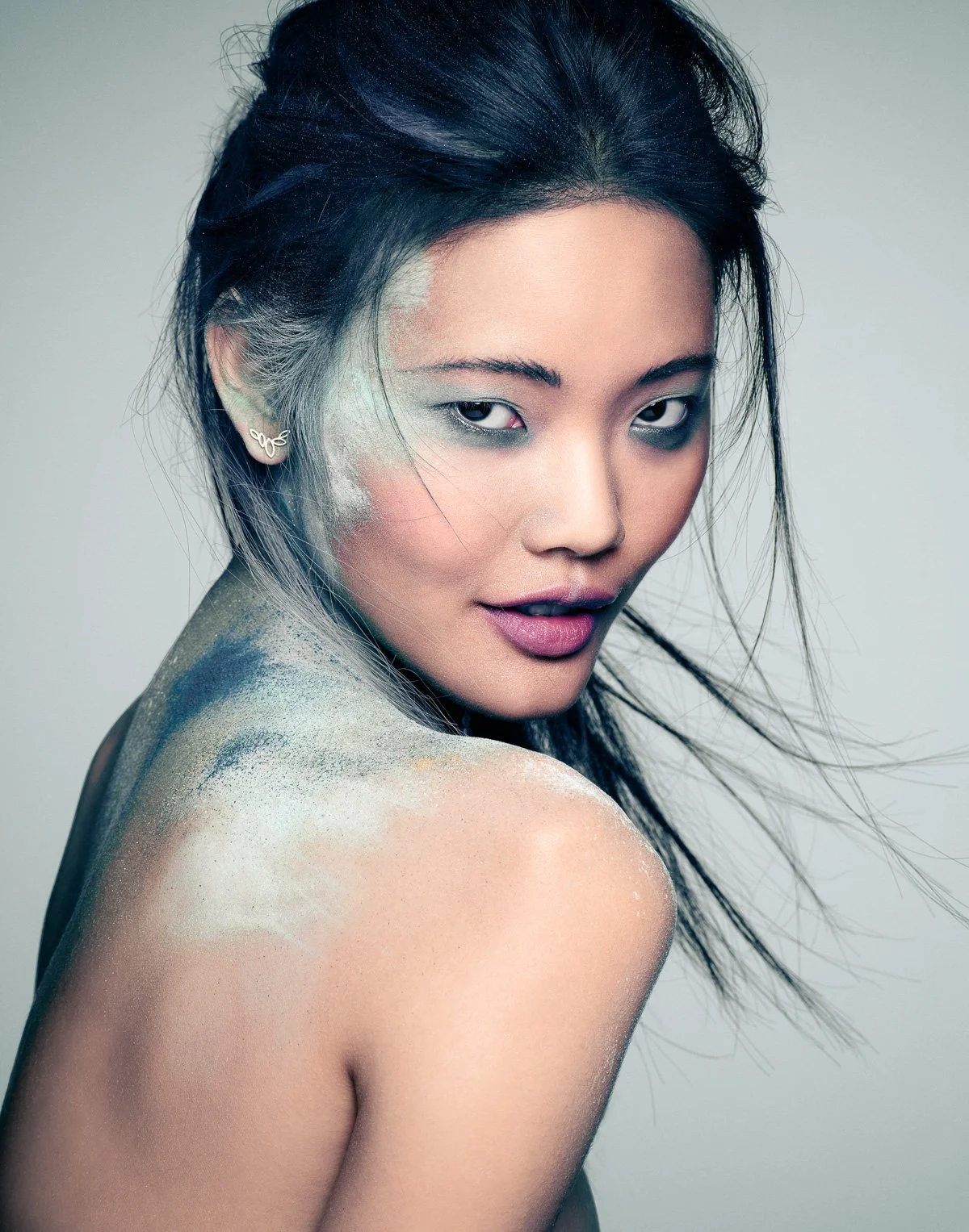 A woman with black hair and dyed silver highlights, looking at the camera with her shoulders exposed. She has glitter on her shoulder and face, and is wearing small, wing-shaped earrings.
