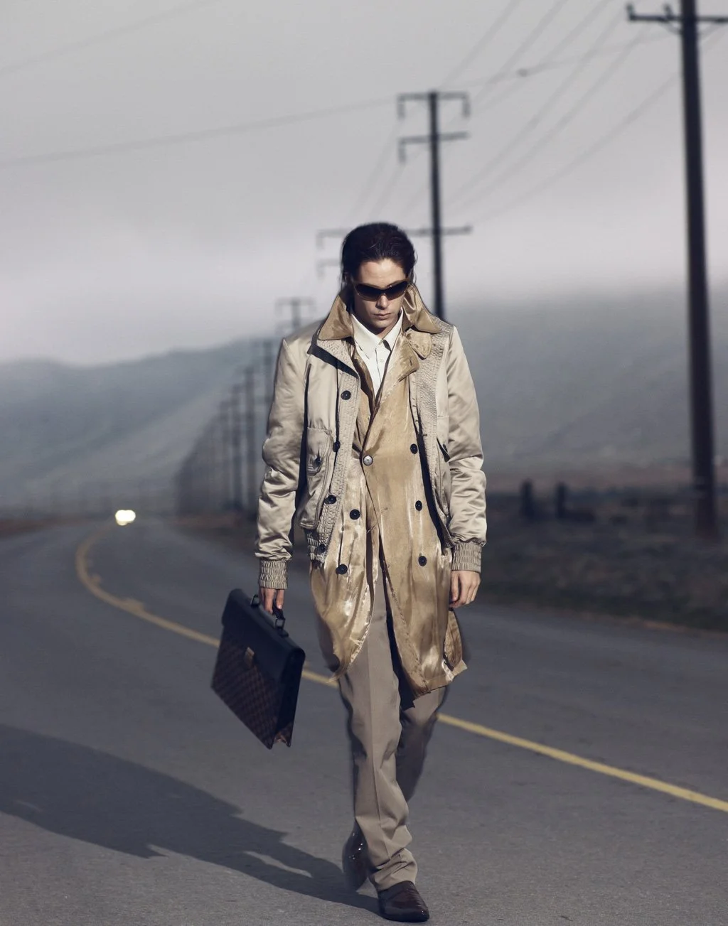 A person with sunglasses walking on a rural road, carrying a black briefcase, wearing a beige/khaki coat and pants, with a background of power lines and overcast sky.