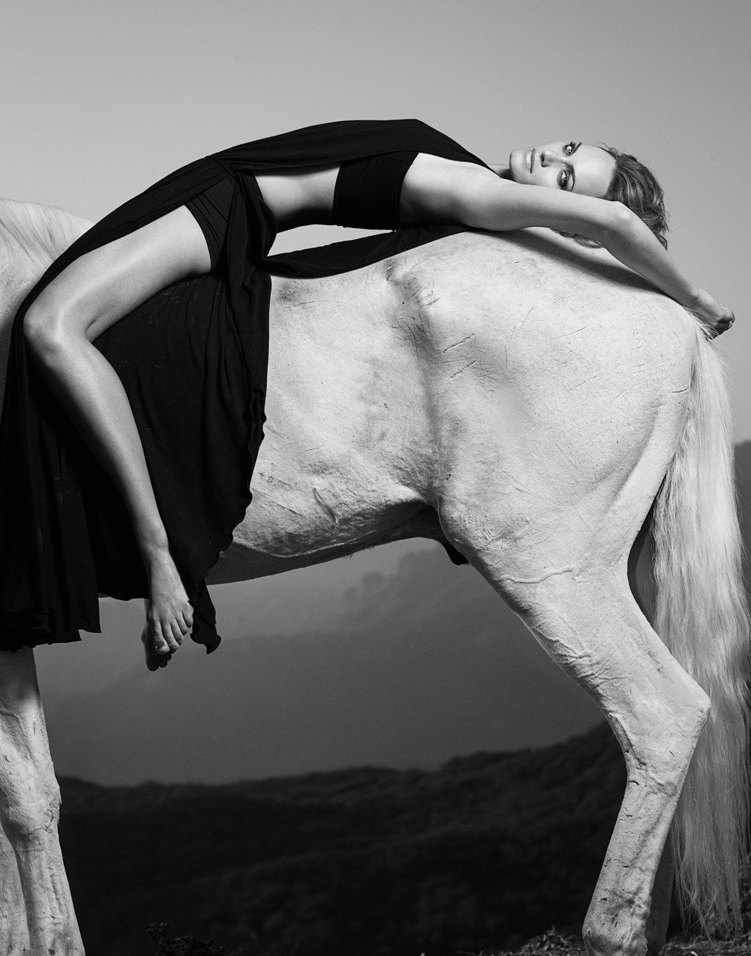 Black and white photo of a woman lying on a large horse with her arms extended over the horse's back, her head resting on her arms, wearing a long black dress.