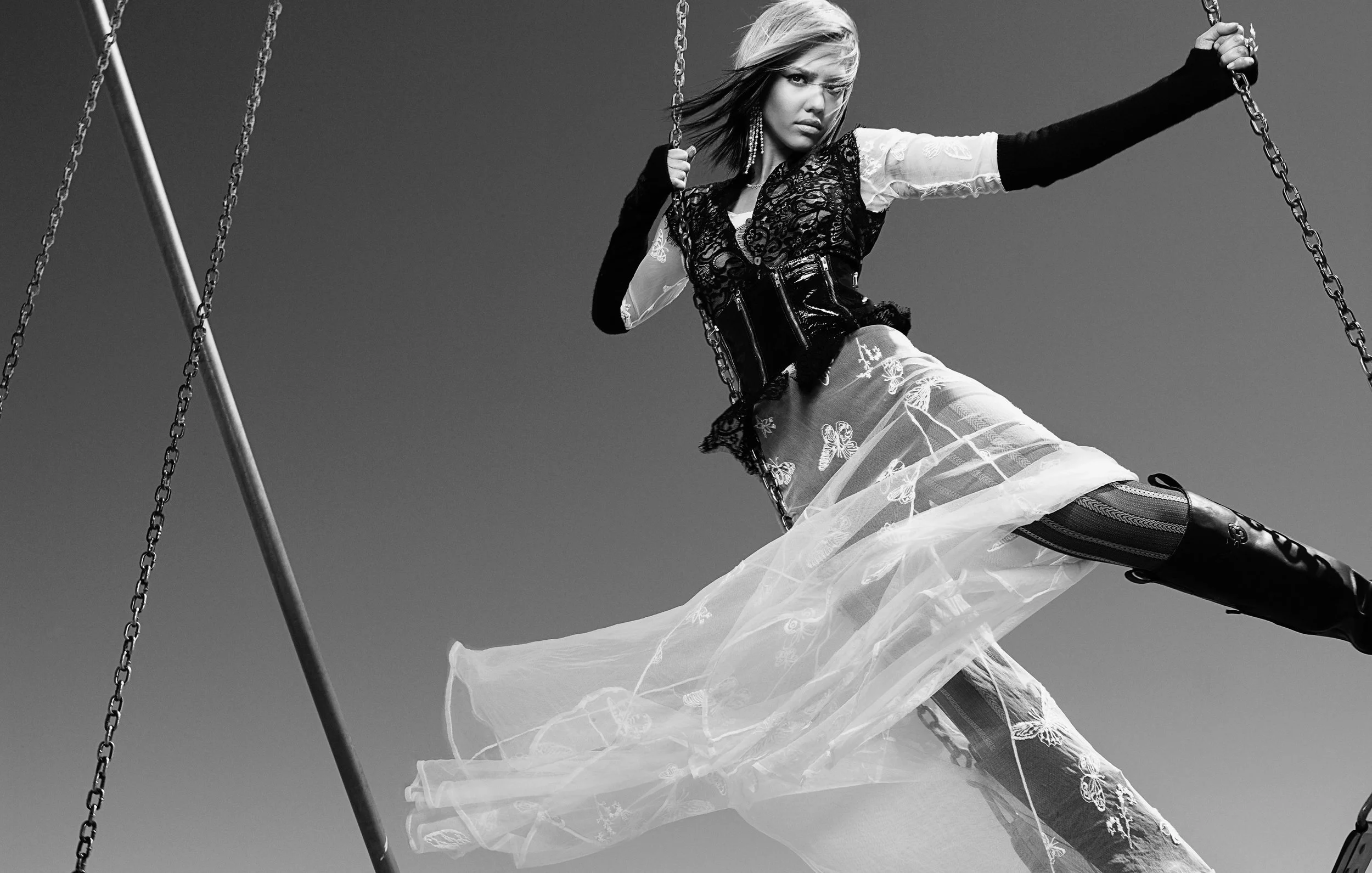 A black and white photo of JESSICA ALBA sitting on a swing, wearing a sheer dress with butterfly embroidery and thigh-high boots, against a plain sky background.