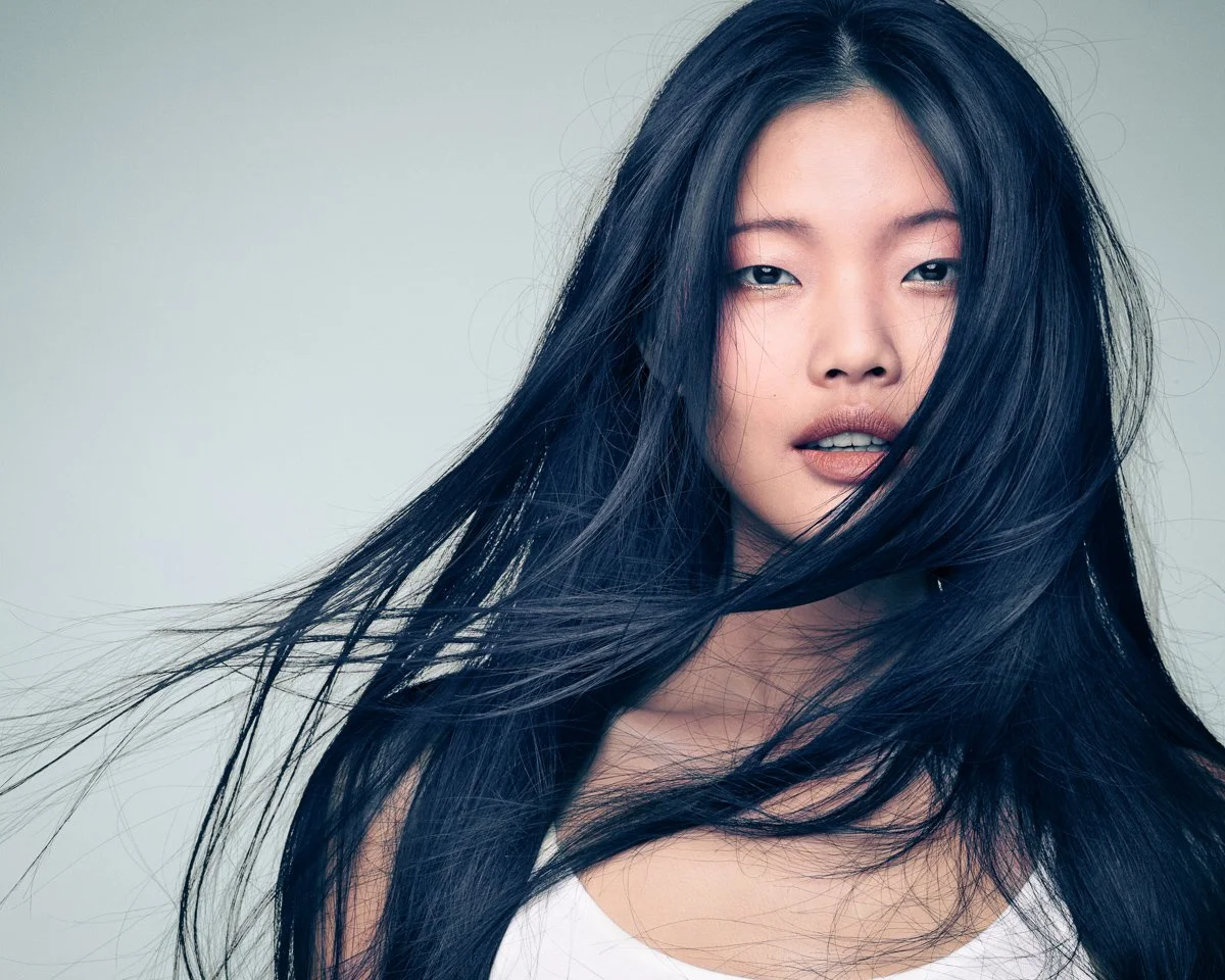 A young woman with long black hair and fair skin, looking at the camera with her hair blowing across her face, against a plain light gray background.