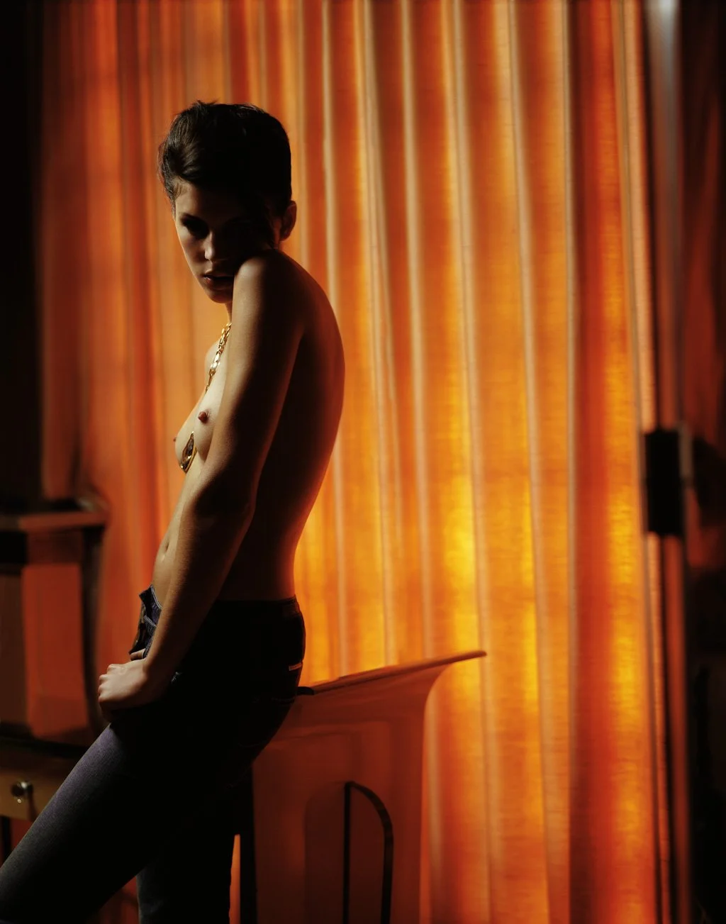 A woman standing near orange curtains, partly shadowed, with short dark hair and wearing a gold chain necklace and pants.