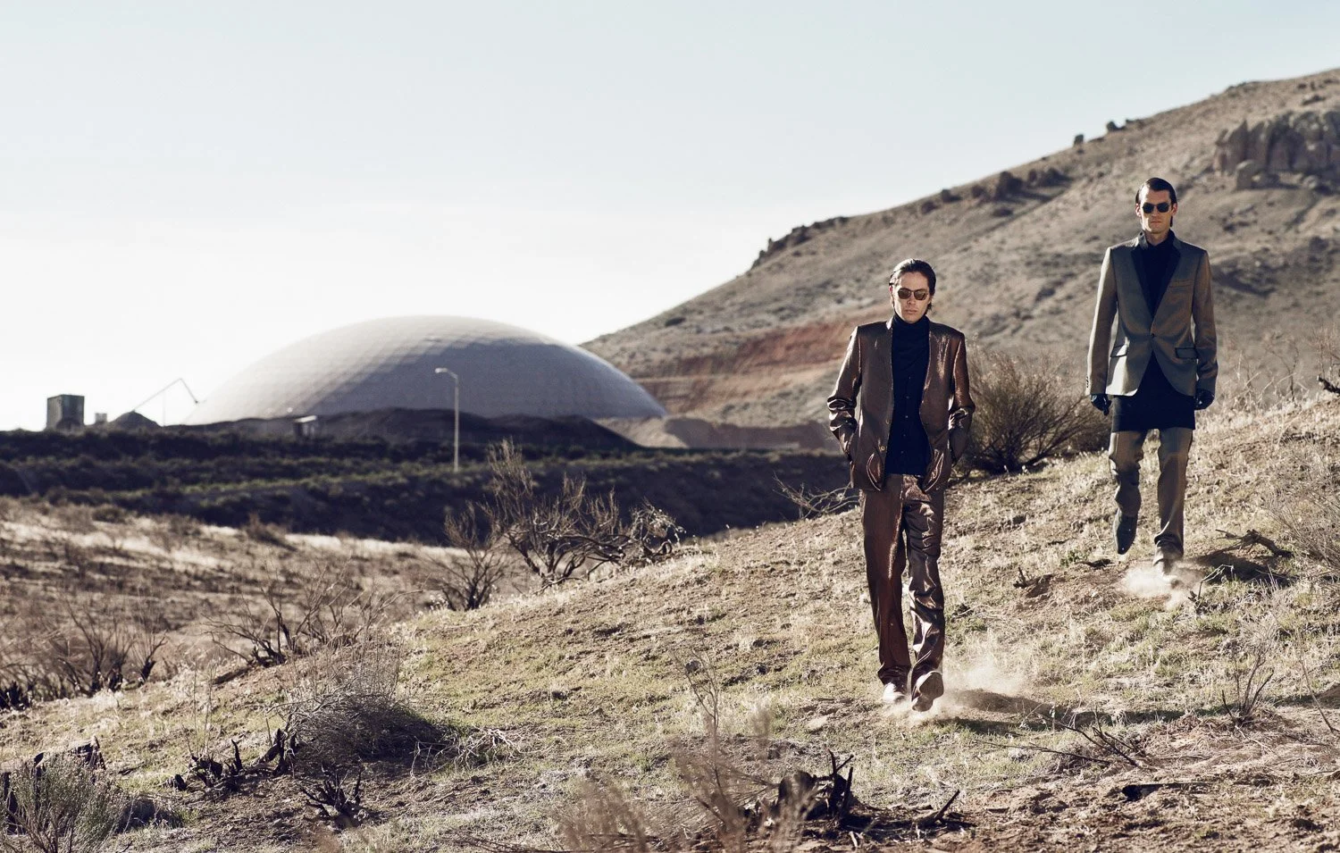Two people walking on a sloped desert landscape with a modern white dome structure in the background, both wearing stylish coats and sunglasses.