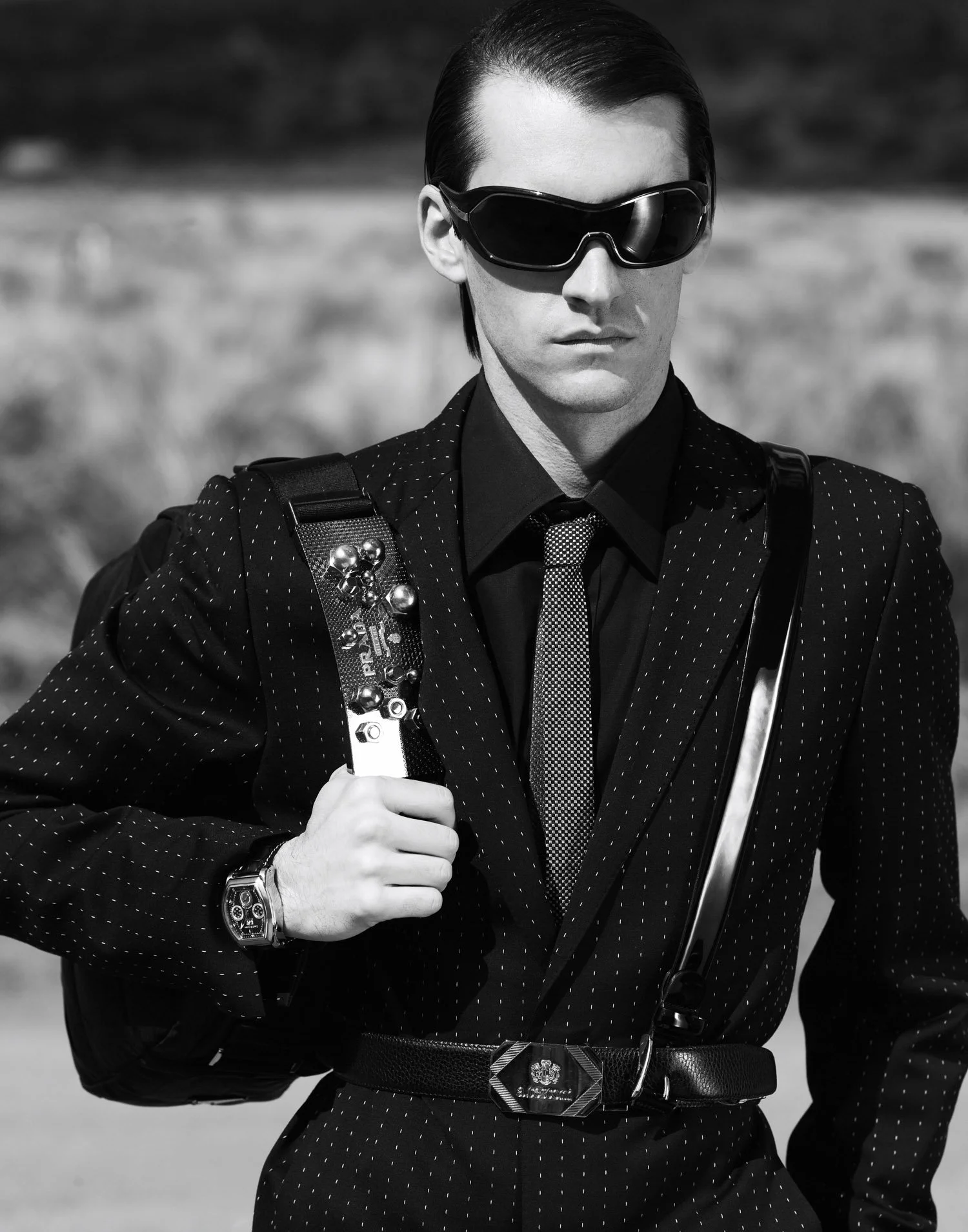 Black and white photo of a man wearing sunglasses, a polka dot suit, and a tie, carrying a backpack and a guitar, outdoors with blurred background.