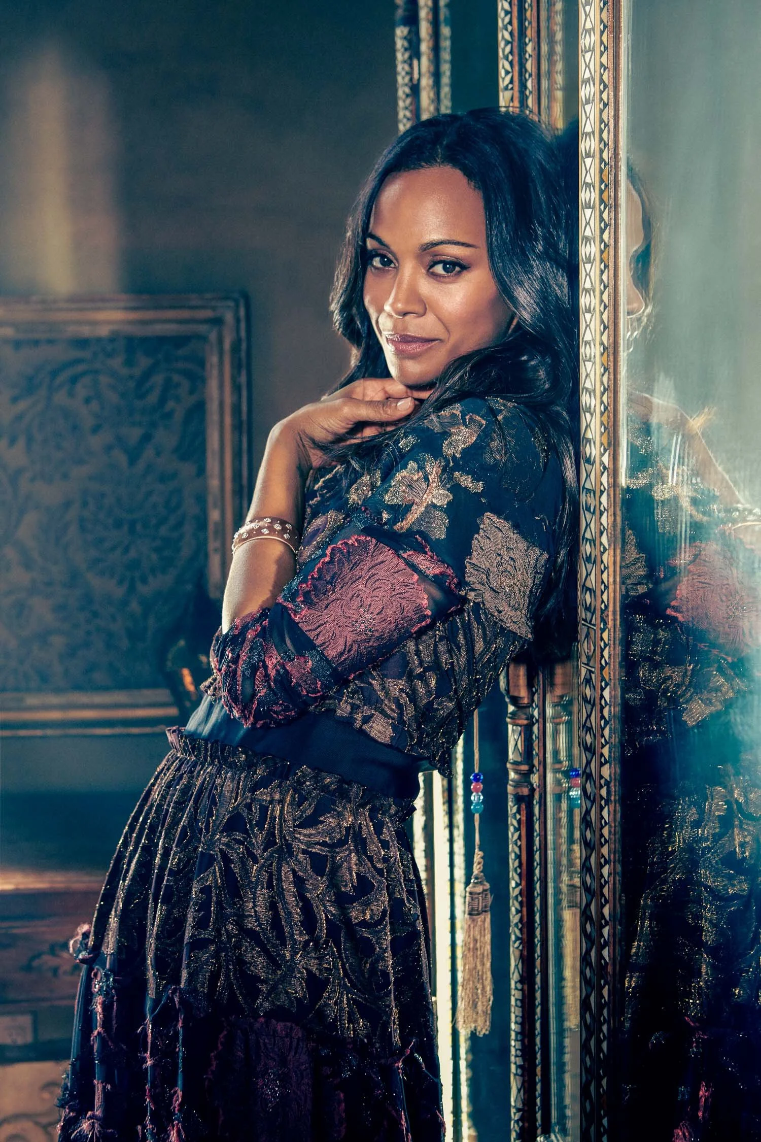 A woman with dark hair standing and touching a mirror, wearing patterned clothing, in a room with ornate decor.